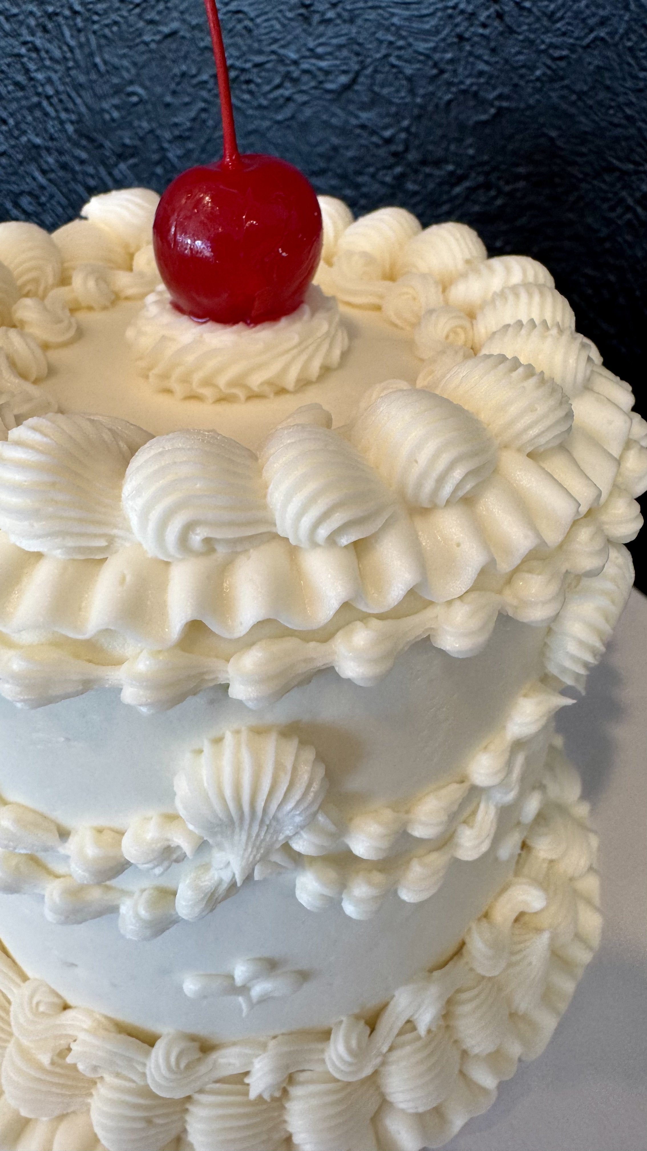 White layered cake decorated with white frosting shells and a single cherry on top.