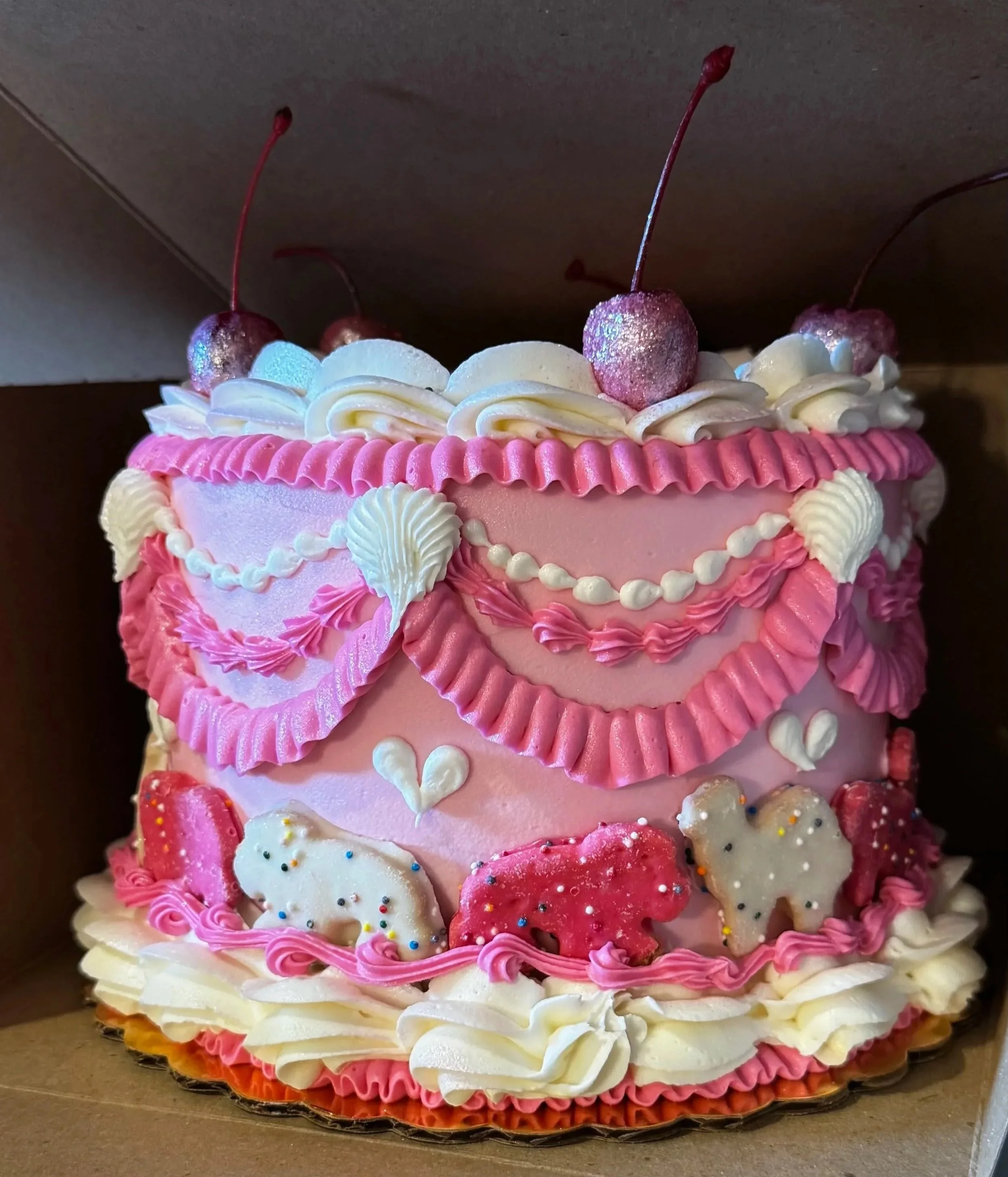 A pink birthday cake decorated with white and pink icing, featuring heart and animal-shaped cookies, cherries on top, and pink and white frosting decorations.
