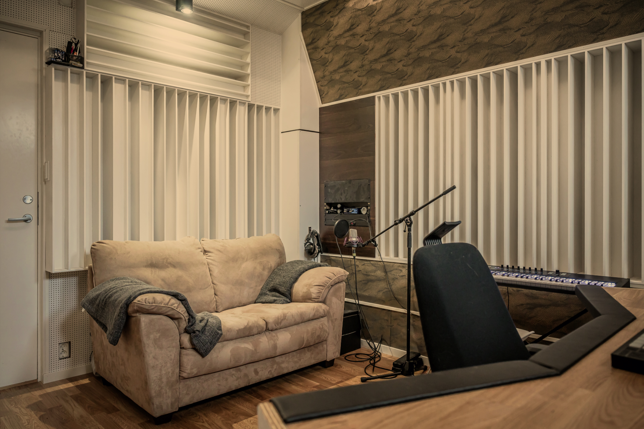 A recording studio room with a beige sofa, black chairs, microphone, and audio equipment. Acoustic panels and a textured ceiling are visible.