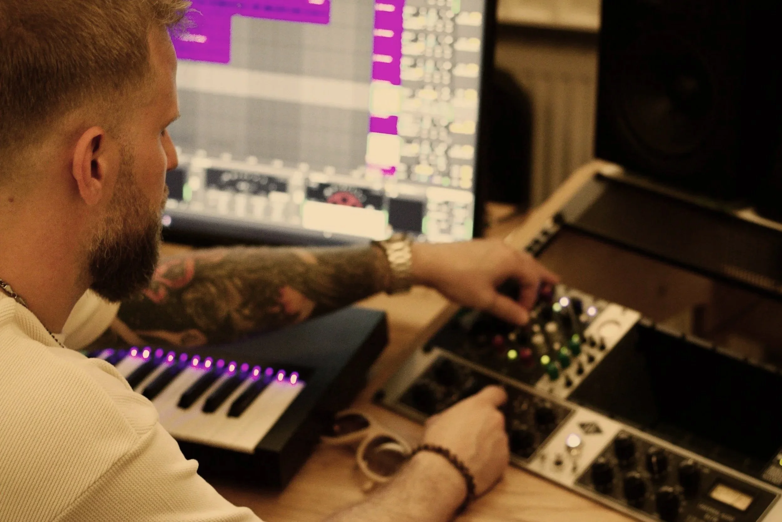 A man with tattoos on his arm working in a music studio with electronic music equipment, including a computer monitor displaying a digital audio workstation, a keyboard synthesizer, and an audio mixer.