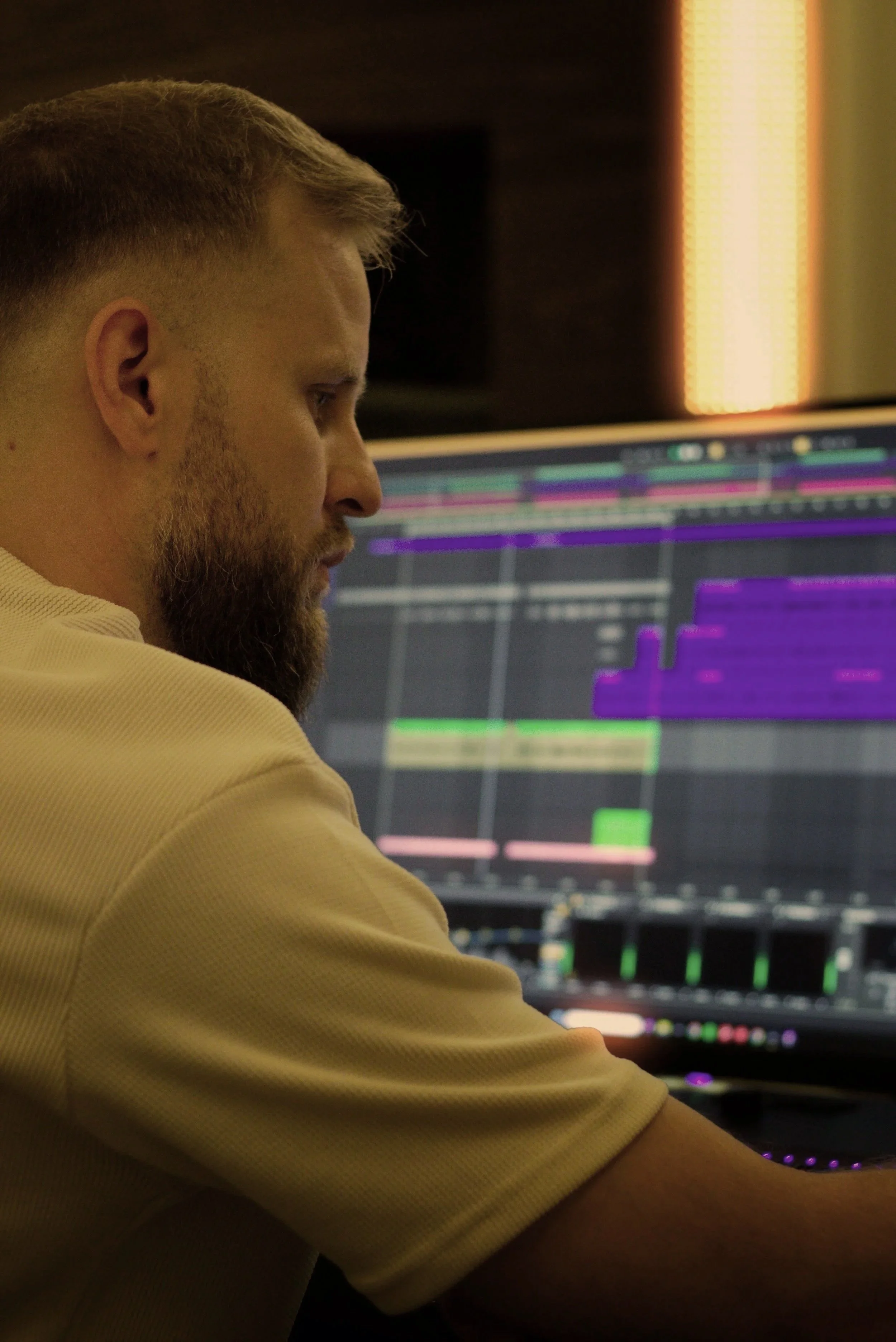 A man with short hair and a beard working on a computer with a large monitor displaying a digital audio workstation.