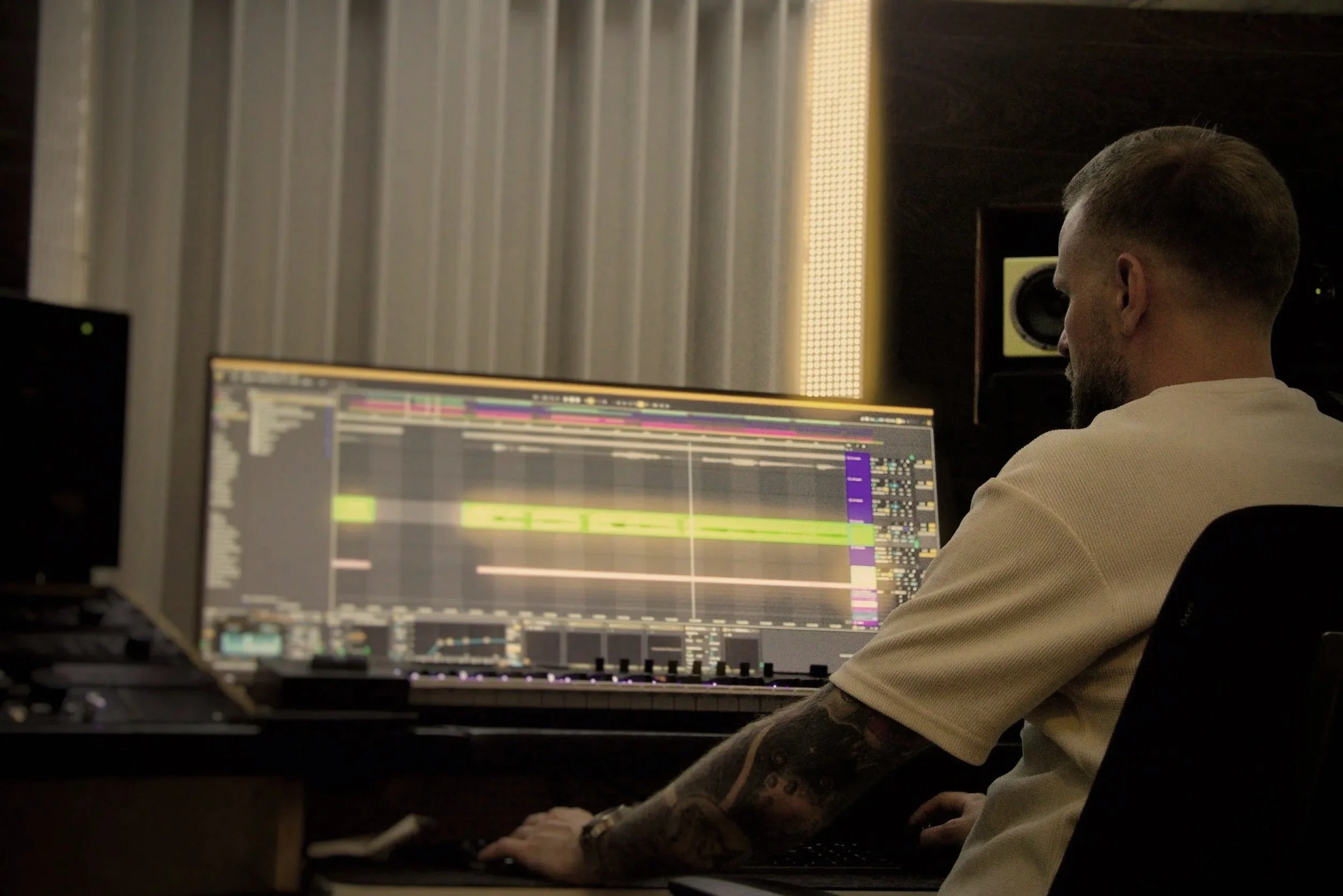 A person with tattoos on their arm using a computer with music production software on the screen in a dimly lit room.