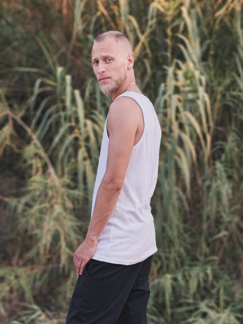 A man with a beard and short hair, wearing a white sleeveless shirt and black shorts, standing outdoors near tall grasses or plants, looking over his shoulder.