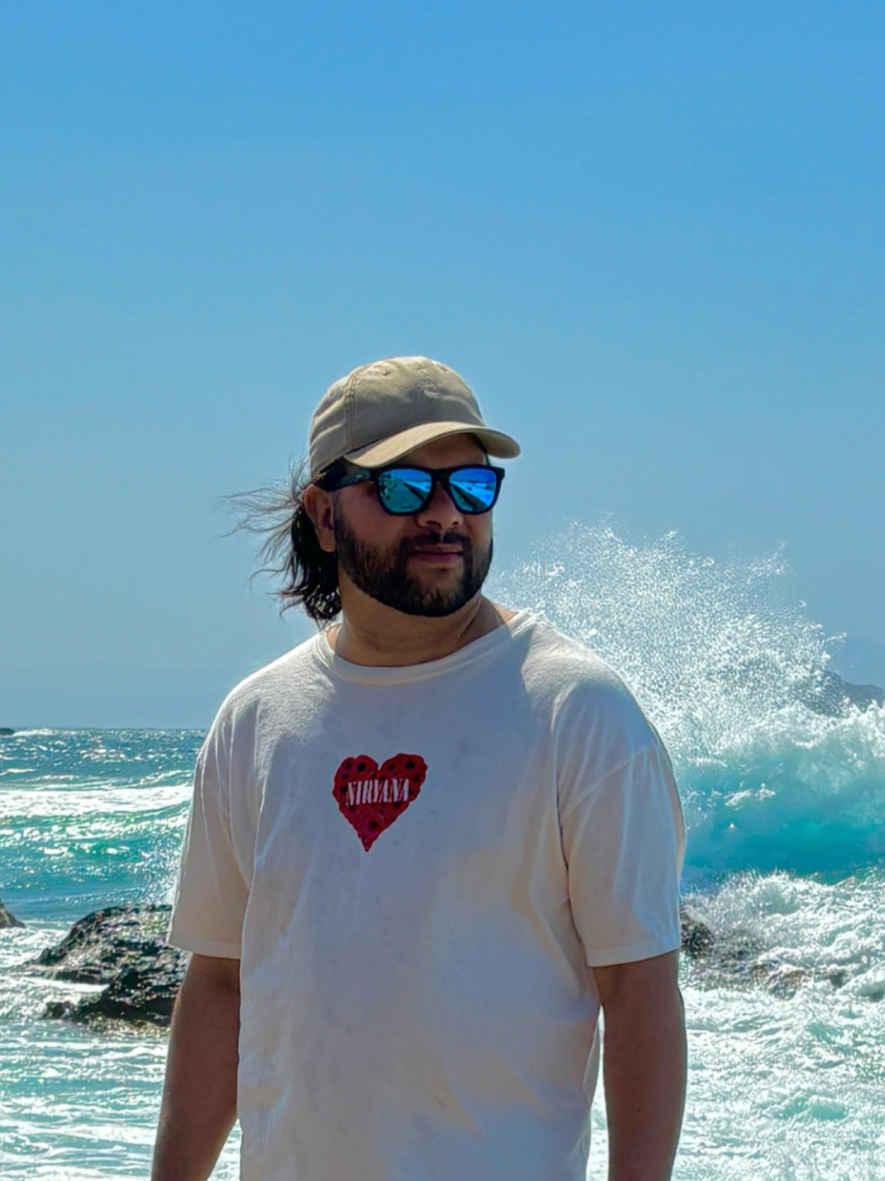 A man with long hair and a beard wearing sunglasses, a beige cap, and a white T-shirt with a red heart graphic, standing by the ocean with waves crashing behind him.