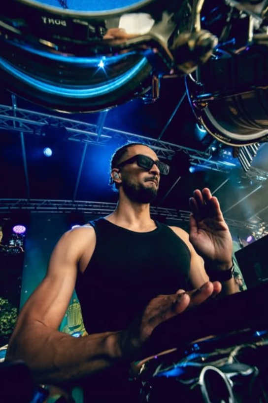 A DJ wearing sunglasses and a black sleeveless shirt performing at a music festival with stage lights and equipment surrounding him.