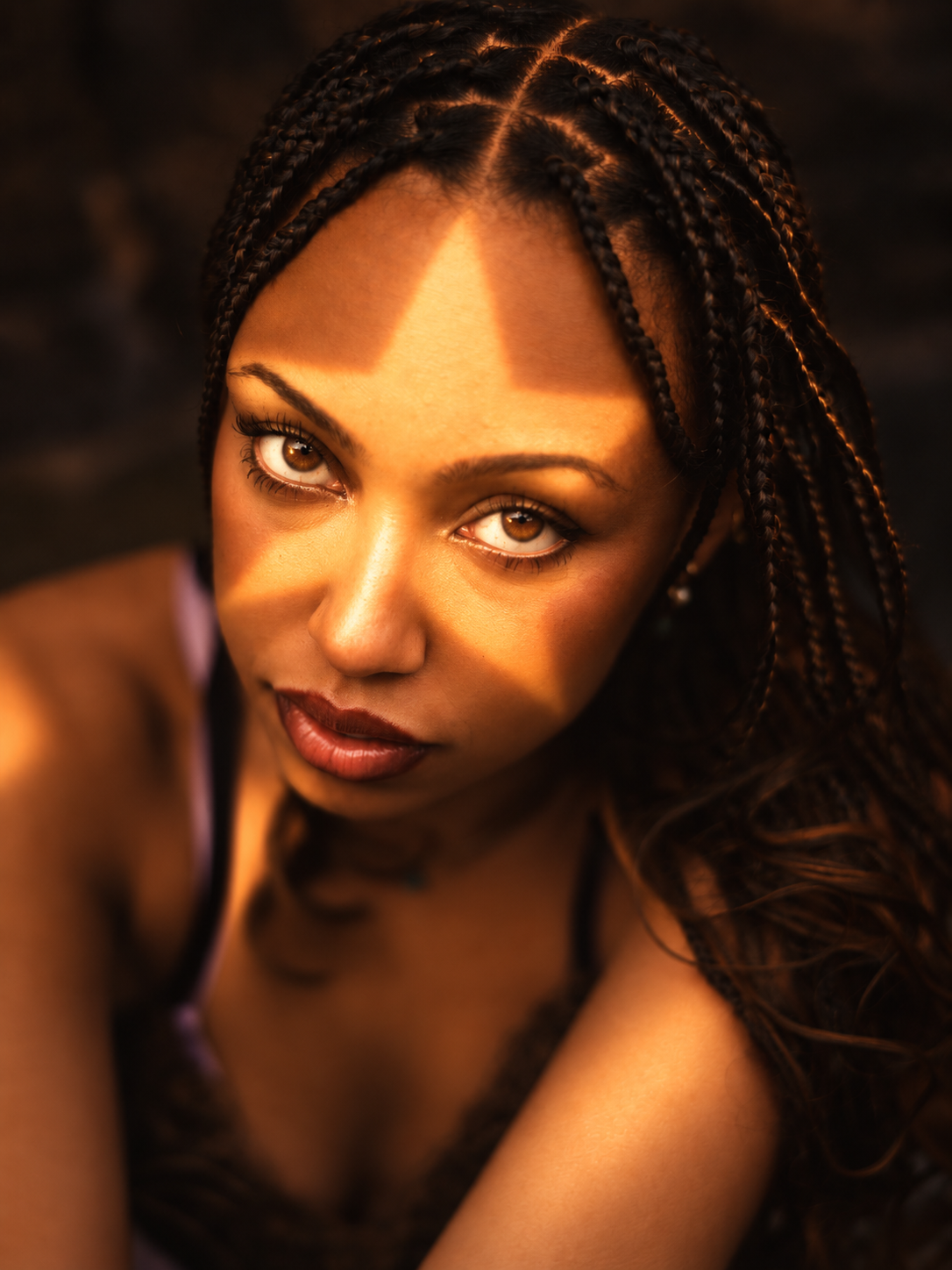 A woman with braided hair and glowing skin, looking directly at the camera with sunlight creating a shadow pattern on her face.