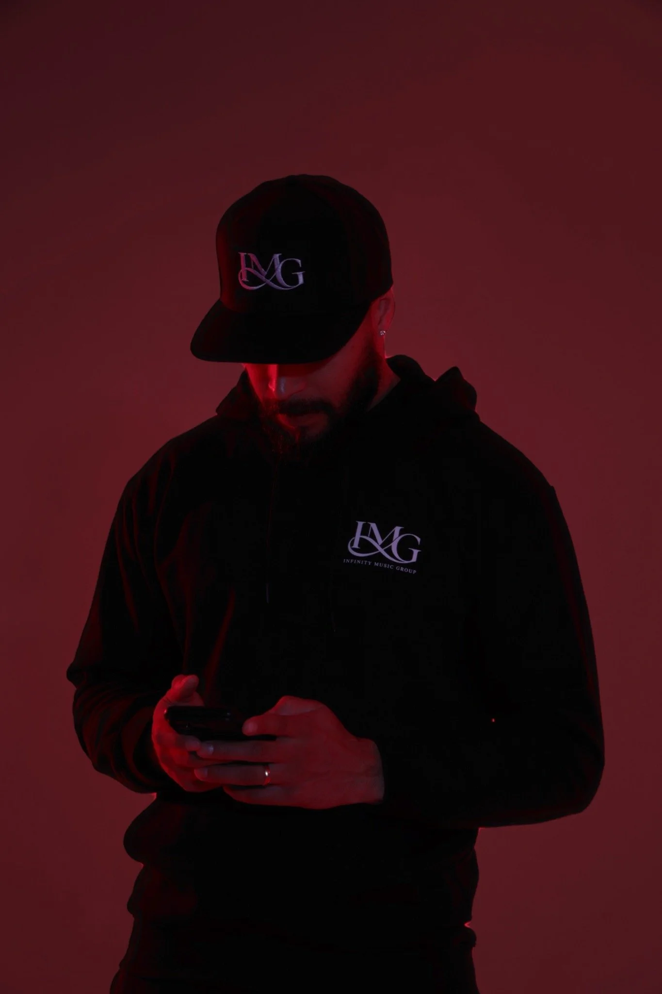 A man wearing a black hoodie and a baseball cap with a logo, looking down at his phone against a red gradient background.