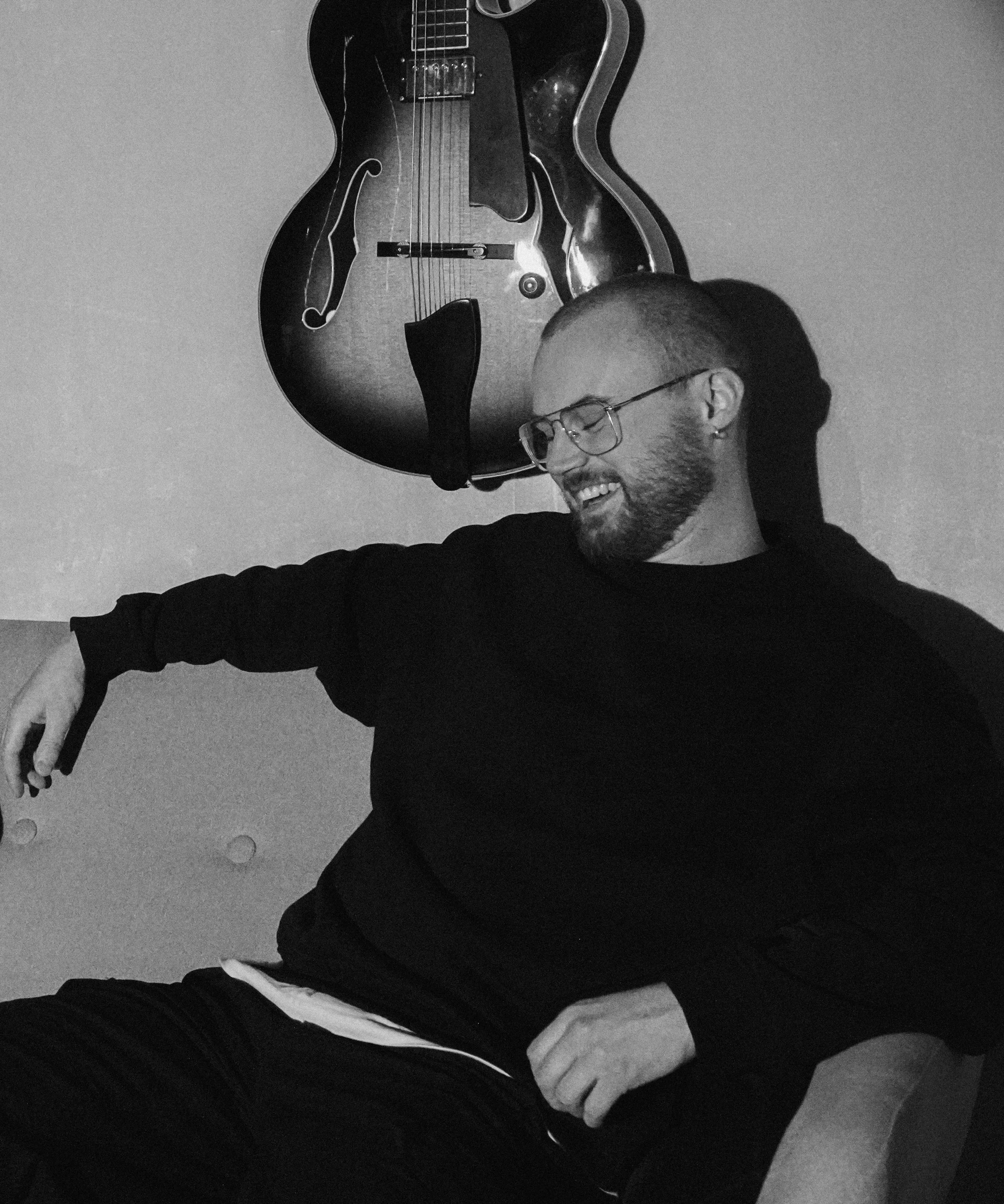 A man with glasses, a beard, and short hair sitting in a music studio. There are three guitars on a stand, an amplifier, and music production equipment on a desk.
