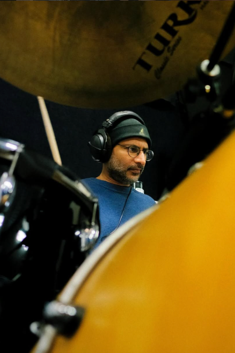 A man with glasses, headphones, and a black beanie hat is seen playing drums, with a yellow acoustic guitar partially visible in the foreground.