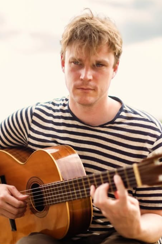 Young man with blond hair wearing a black and white striped shirt, playing an acoustic guitar outdoors.