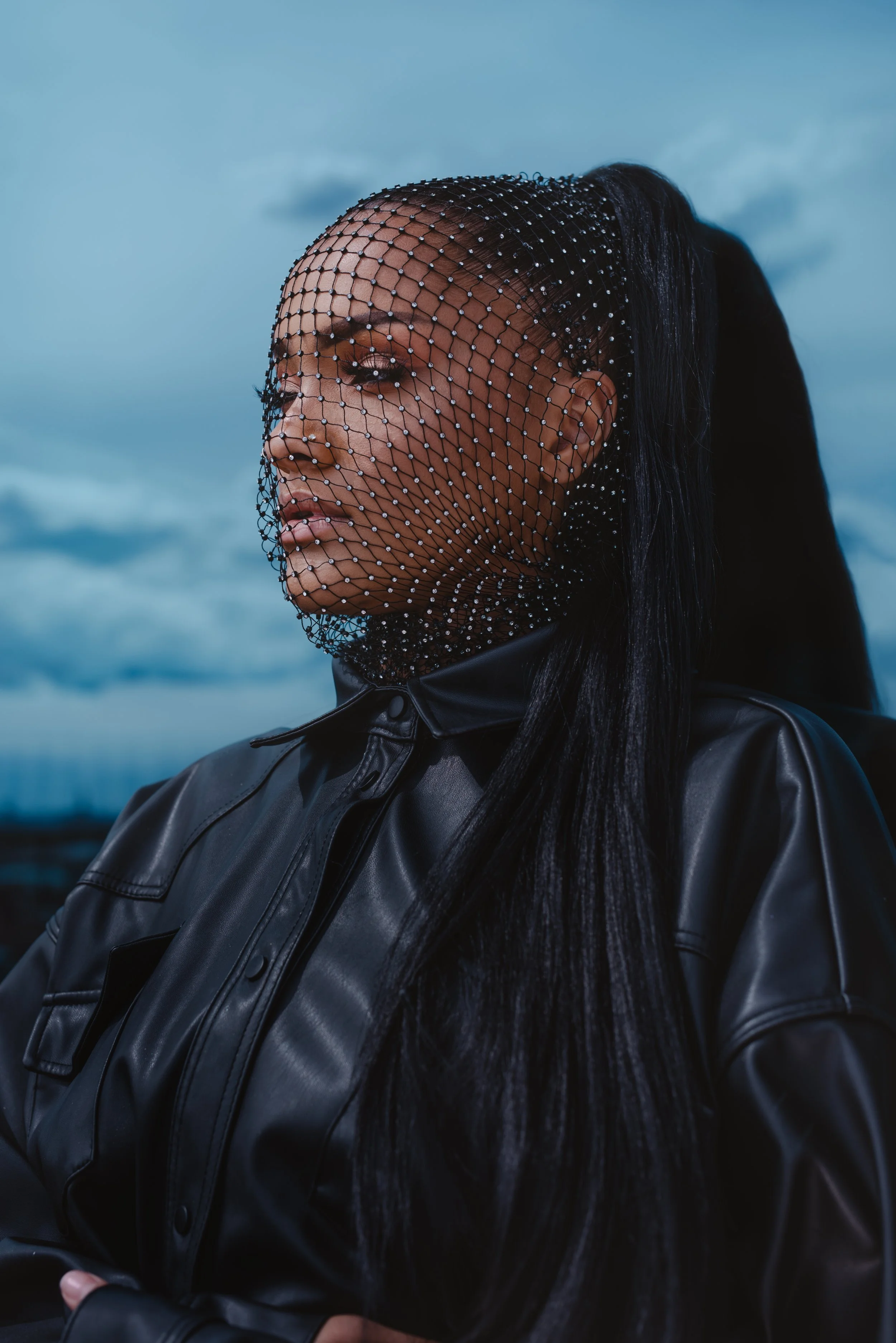 A woman with long dark hair wearing a black leather jacket and a black netted veil adorned with small beads standing outdoors against a blue sky with clouds.