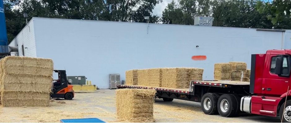 A red flatbed truck carrying hay bales is parked in an outdoor lot, with some hay bales stacked nearby and a forklift moving hay.