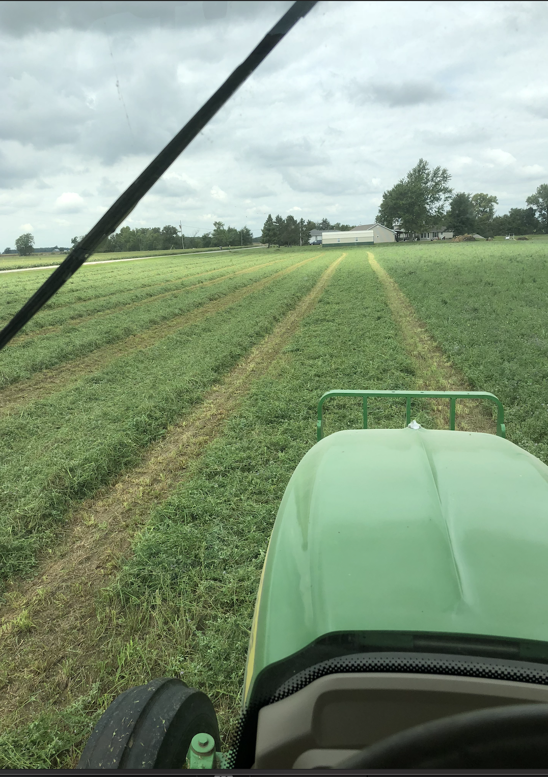 View from inside a tractor cabin showing green fields and a farm in the distance on a cloudy day.