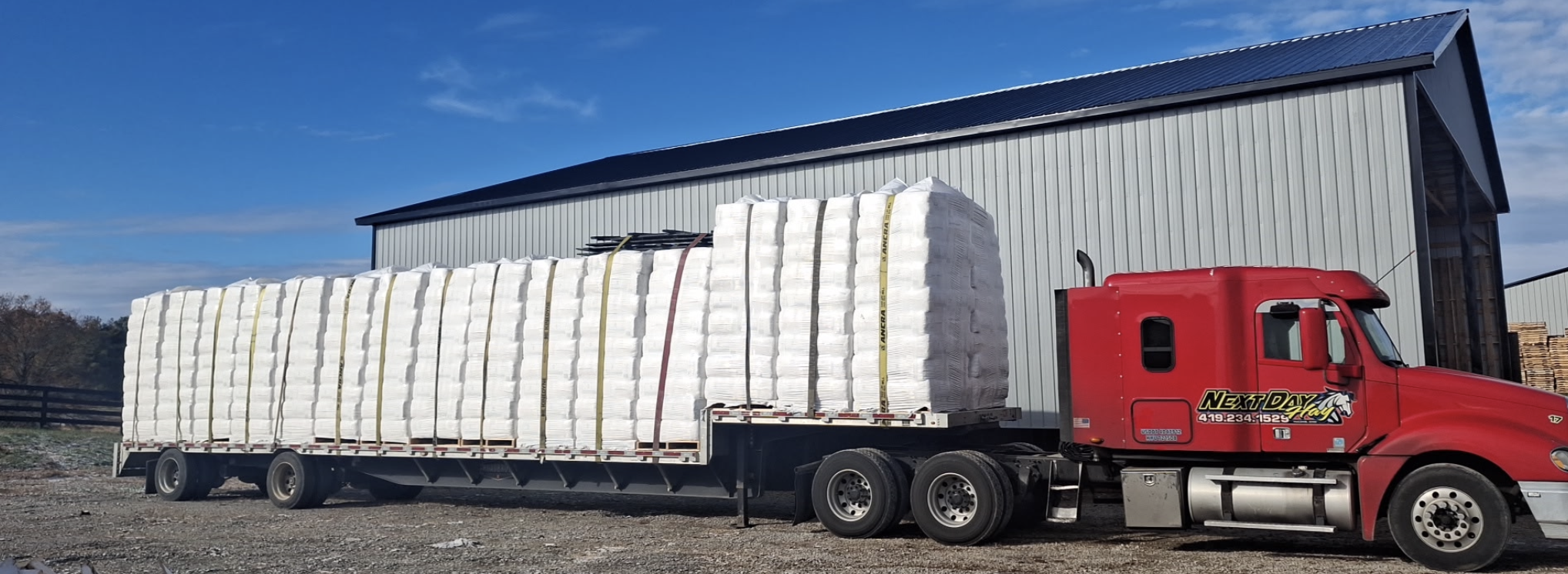 Red semi-truck parked outside a blue metal warehouse, carrying a large load of white rectangular wrapped bales secured with straps.
