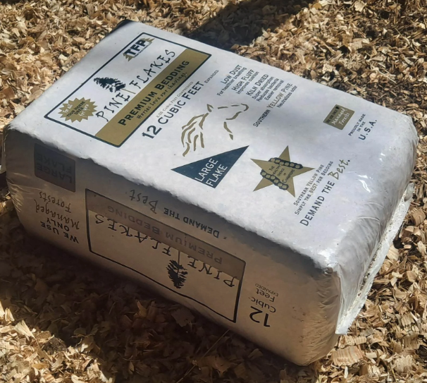 A bag of Pine Flakes premium bedding, 12 cubic feet, intended for large pet enclosures, lying on wood shavings outdoors.