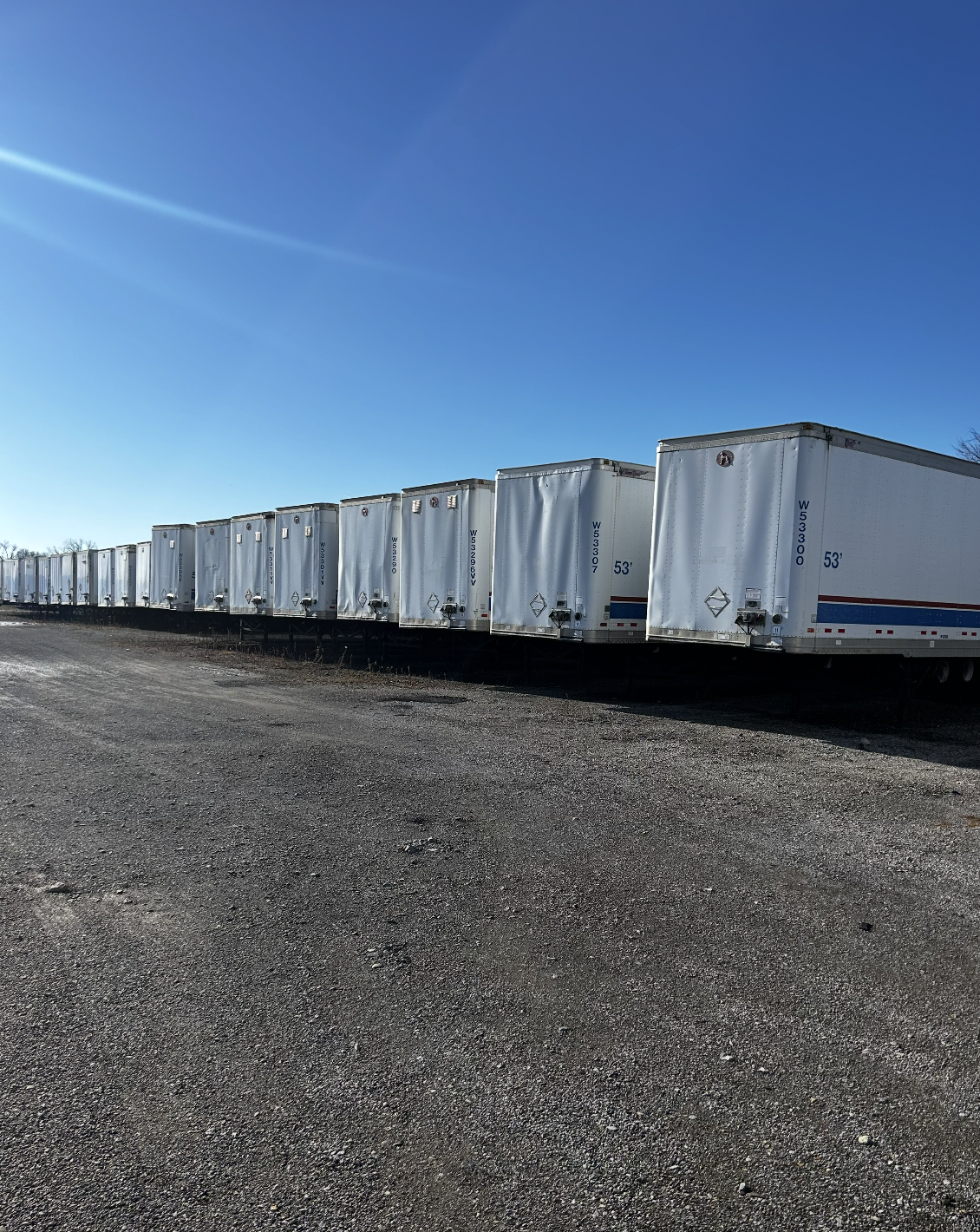 A row of white semi-trailer trucks parked on a gravel lot under a clear blue sky.