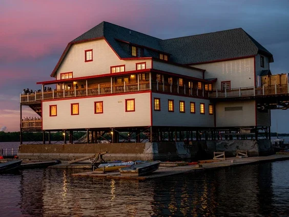 Large house built on stilts over water with a dock and boats, at sunset.