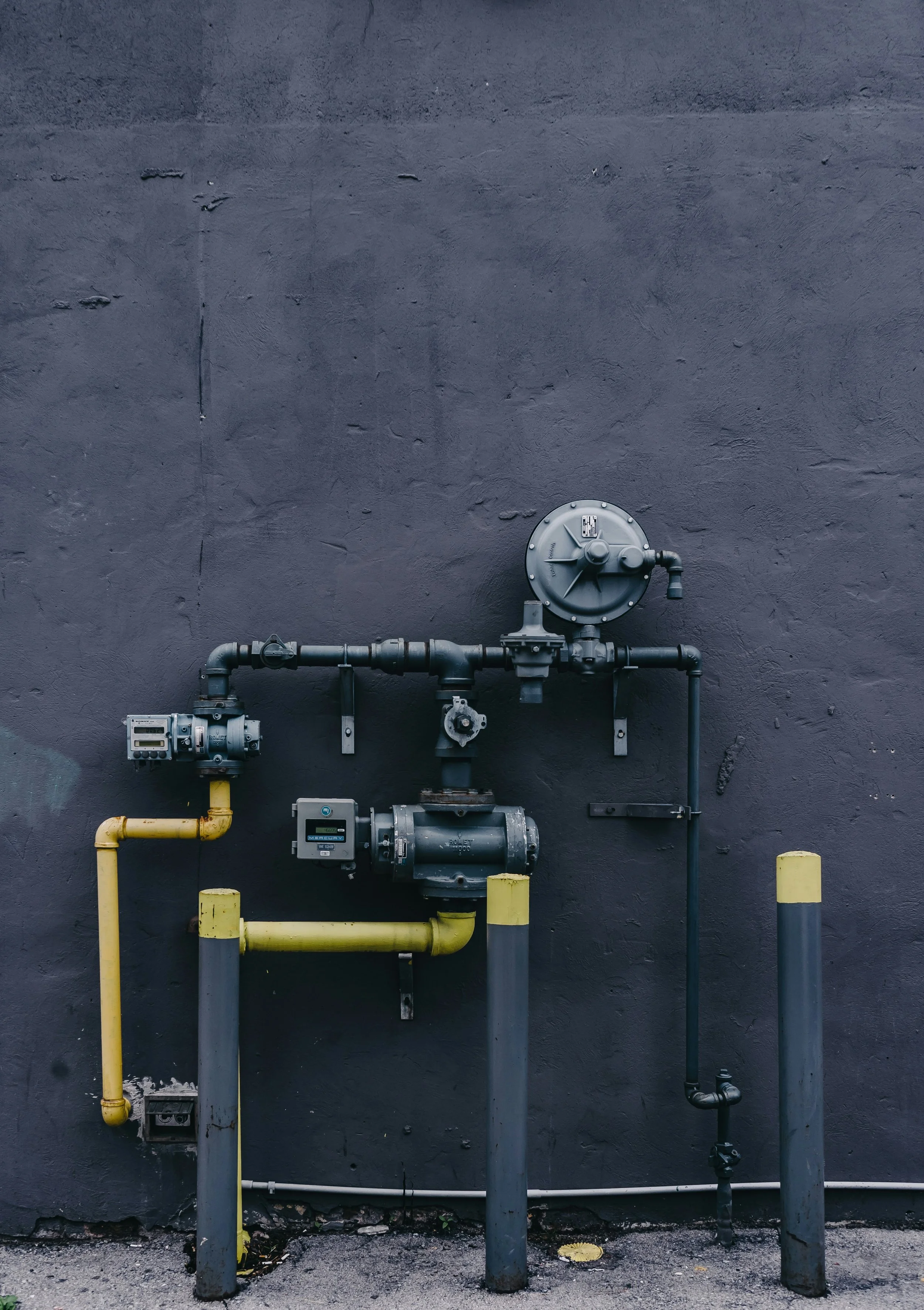 Industrial gas meters and pipes mounted on a dark wall with yellow and gray fixing poles.