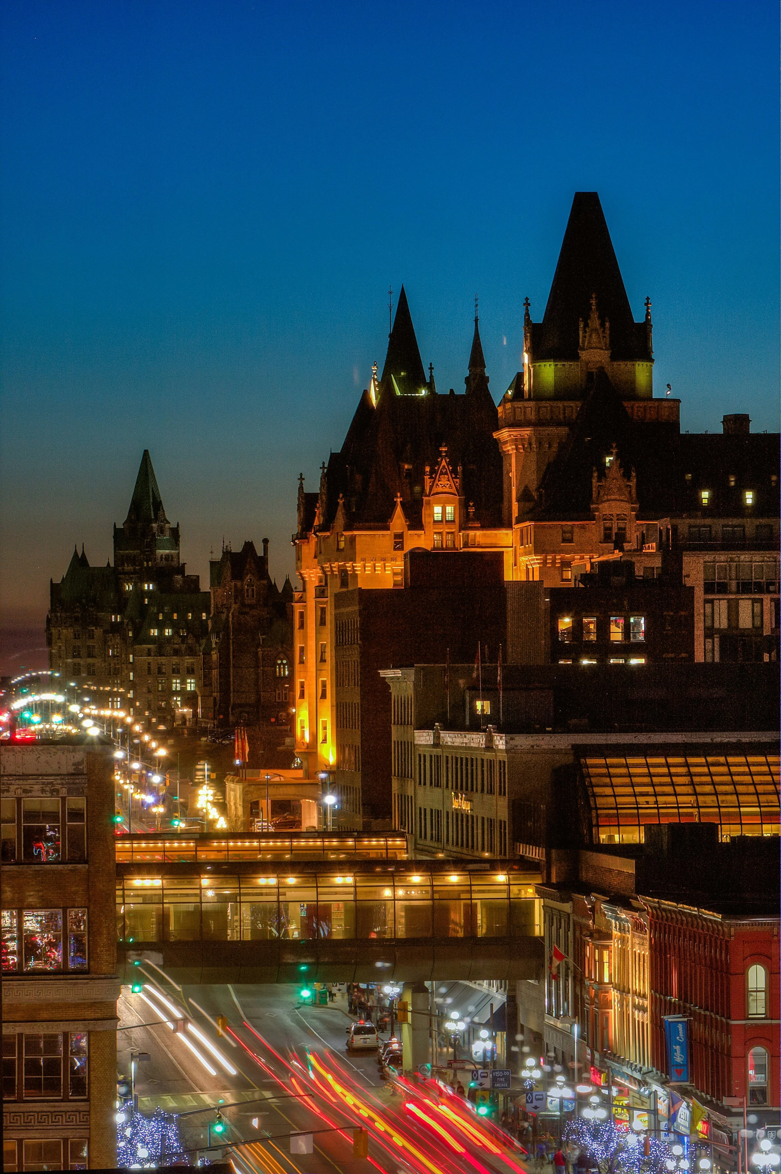 Night view of downtown city with historic castle-like building illuminated, busy street with streaks of car lights, and decorated trees with lights in the foreground.
