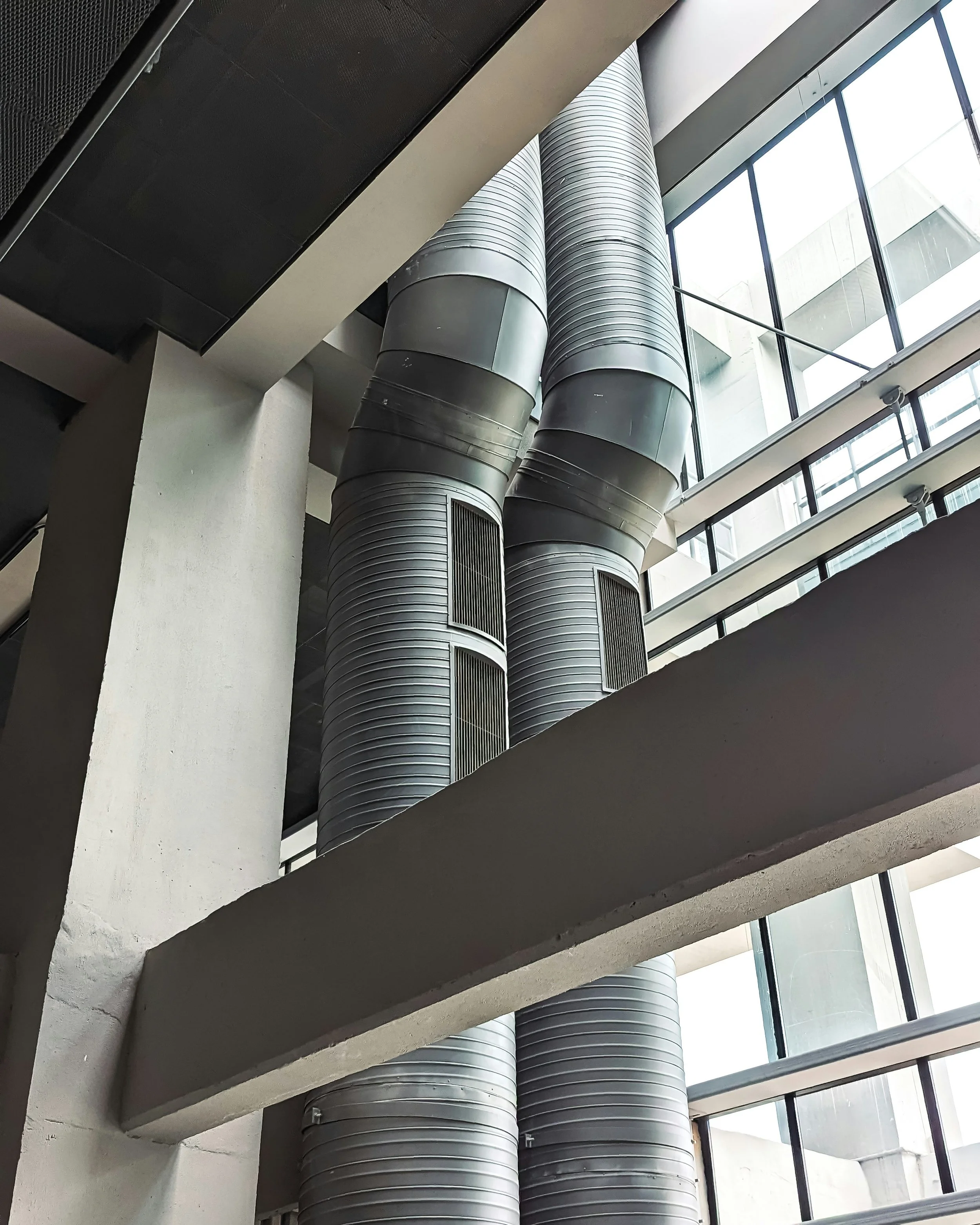 Three large metallic industrial ventilation ducts extend vertically along a wall in a modern building with large glass windows in the background.