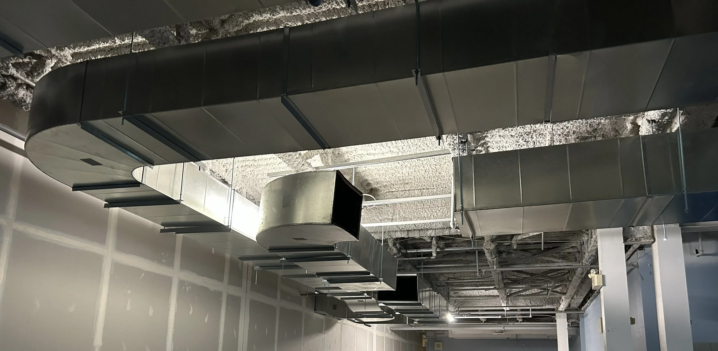 An unfinished commercial kitchen ceiling showing exposed ductwork, insulation, and partially installed drywall, with suspended black duct components.