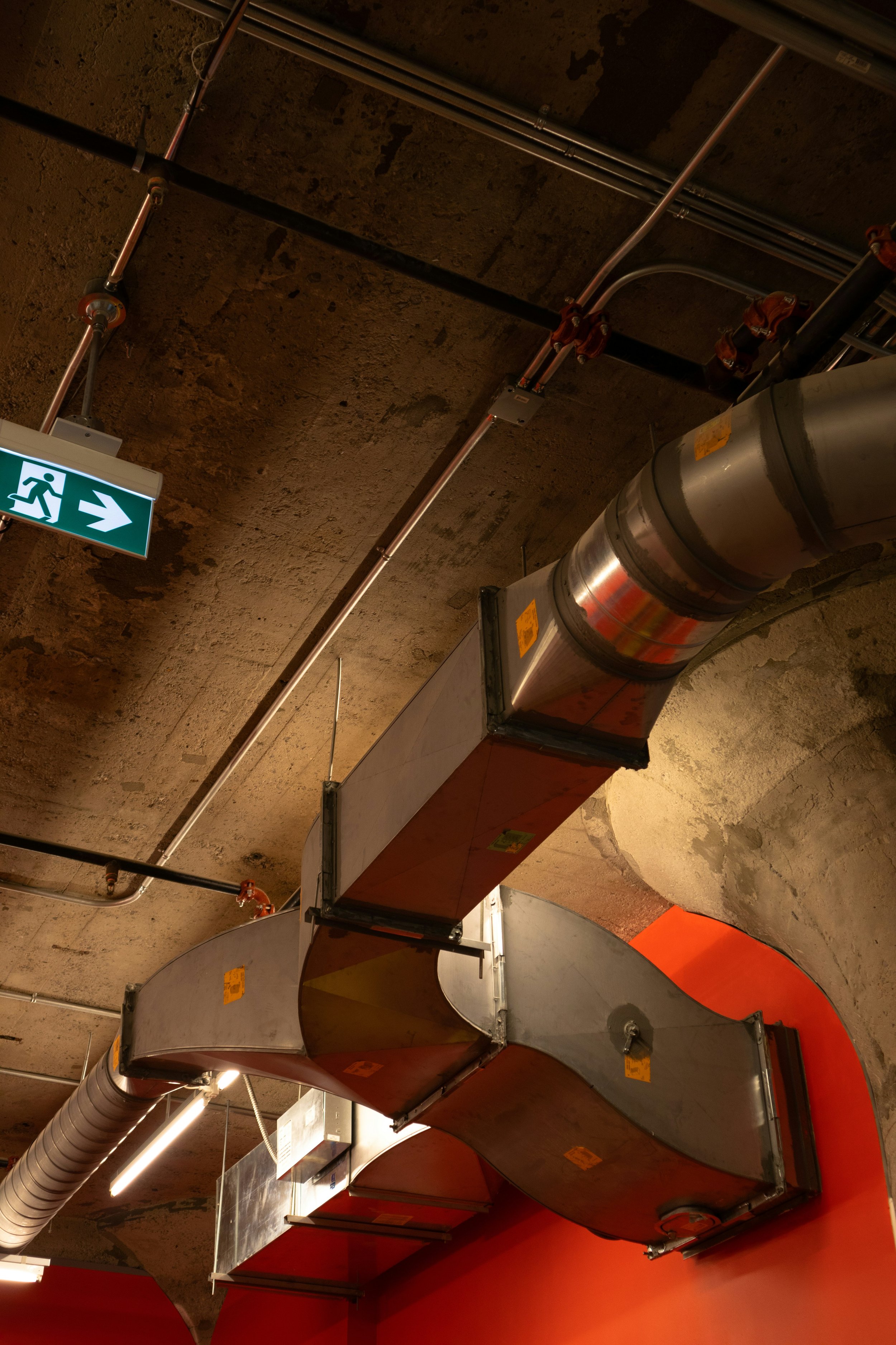 Industrial ceiling with large metal ventilation ducts, pipes, and an emergency exit sign with a running figure and arrow.