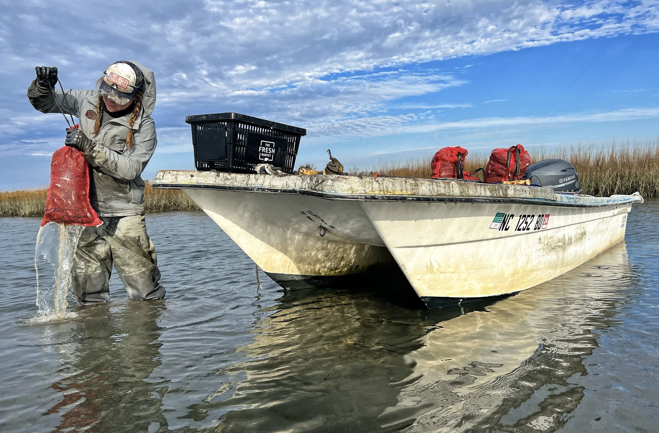 A woman in waders standing in shallow water beside a white boat, holding a red mesh bag with water dripping from it, with a cloudy blue sky and marshland in the background.