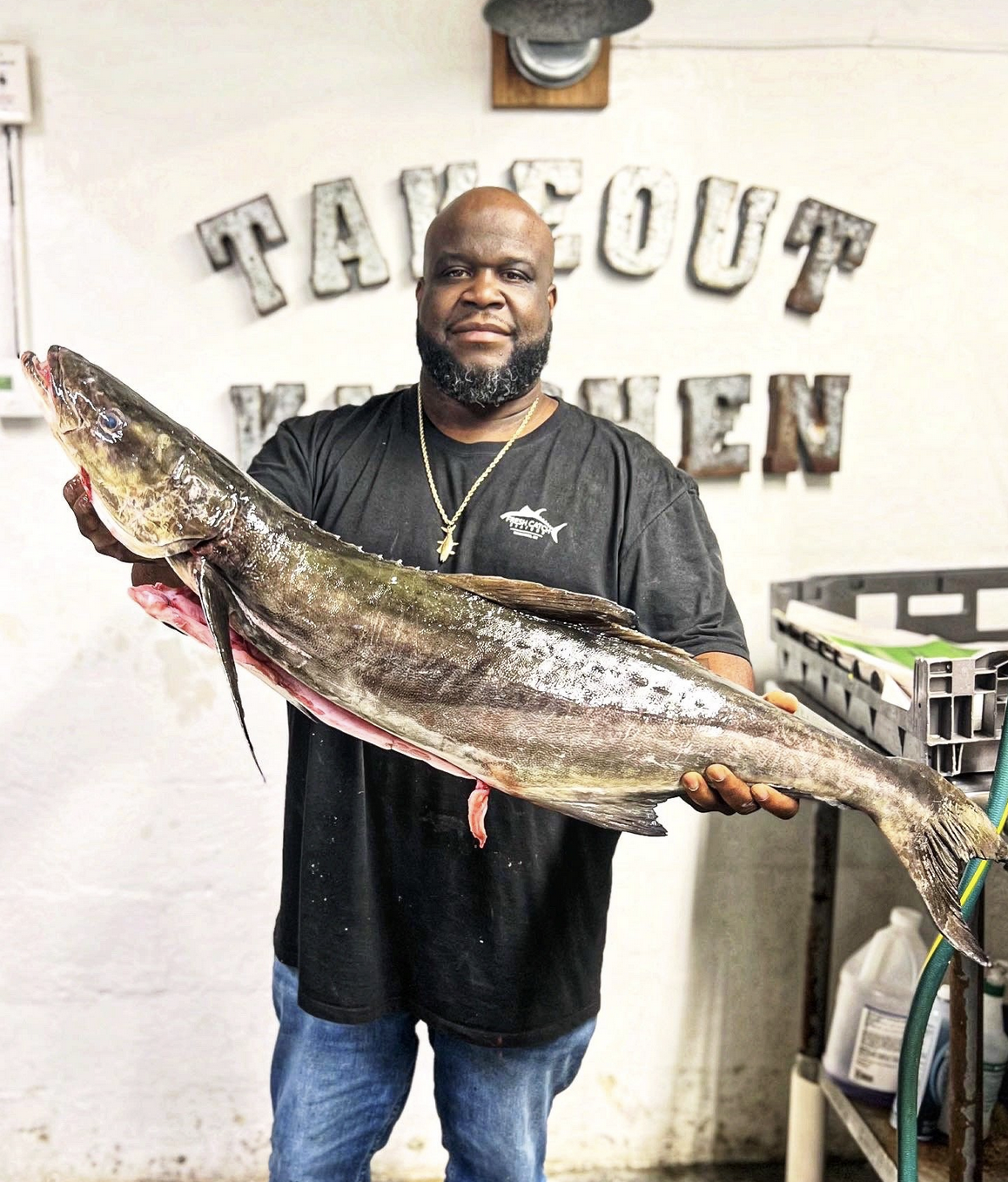 A man with a beard holding a large fish at a seafood market or restaurant, with the sign 'Take Out Kitchen' in the background.