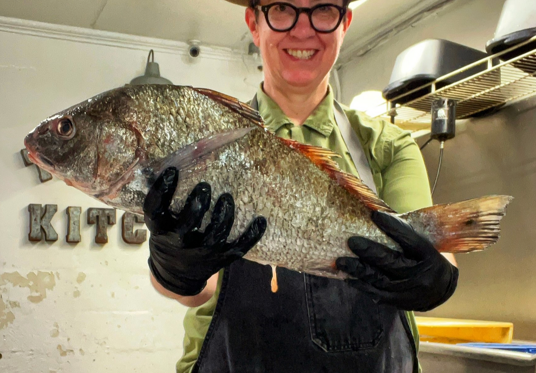 A person wearing glasses and black gloves holding a large fish indoors.