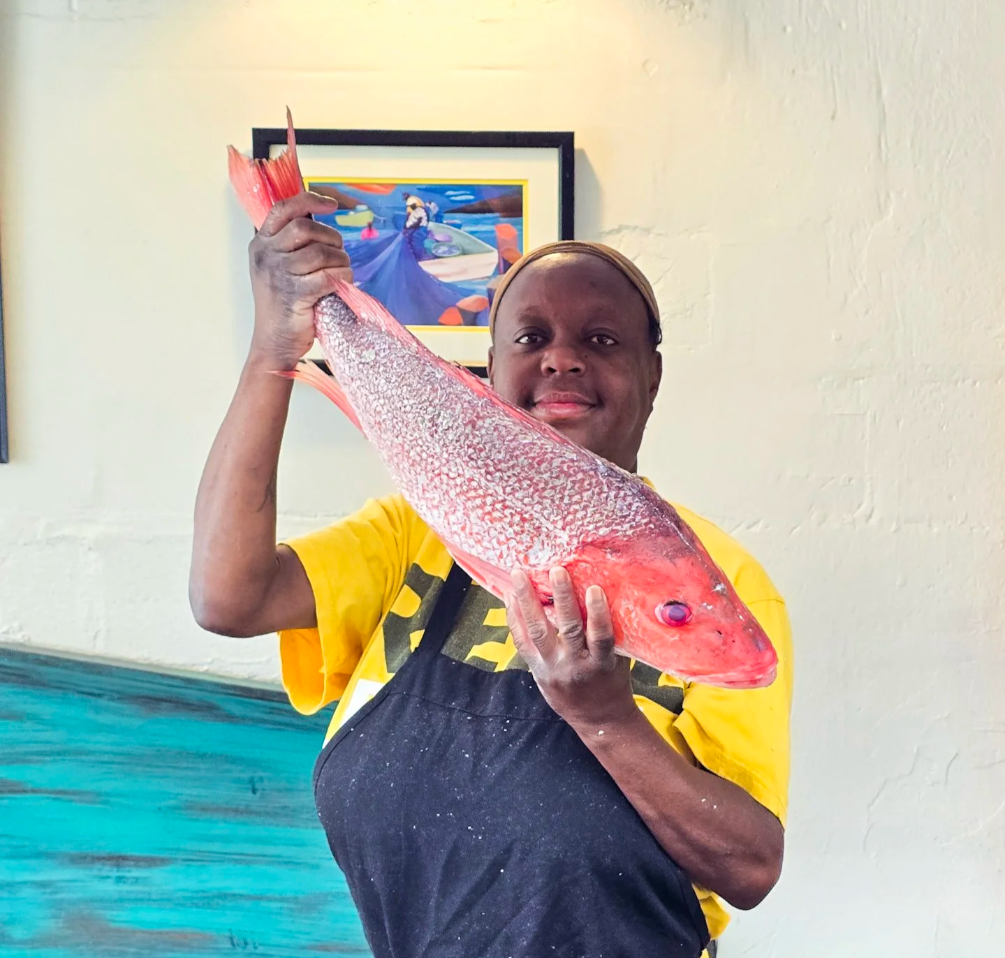 A woman holding a large red fish indoors, with framed art on the wall behind her.