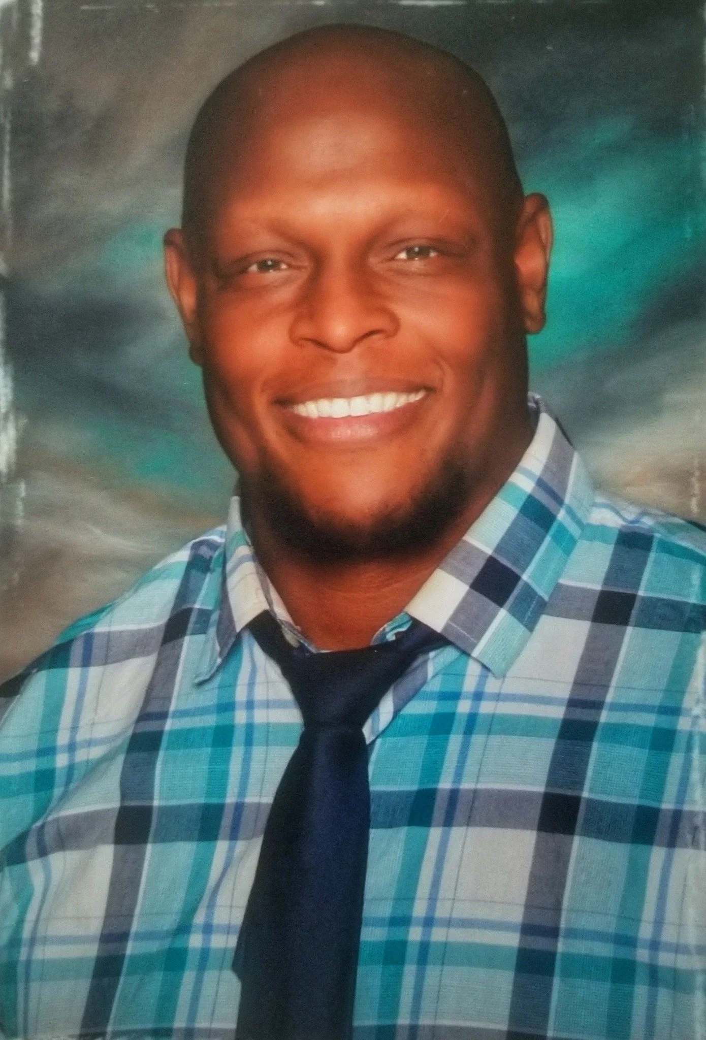 Portrait of a smiling man with a shaved head, wearing a blue, white, and gray plaid shirt and a dark tie, against a colorful, blurred background.
