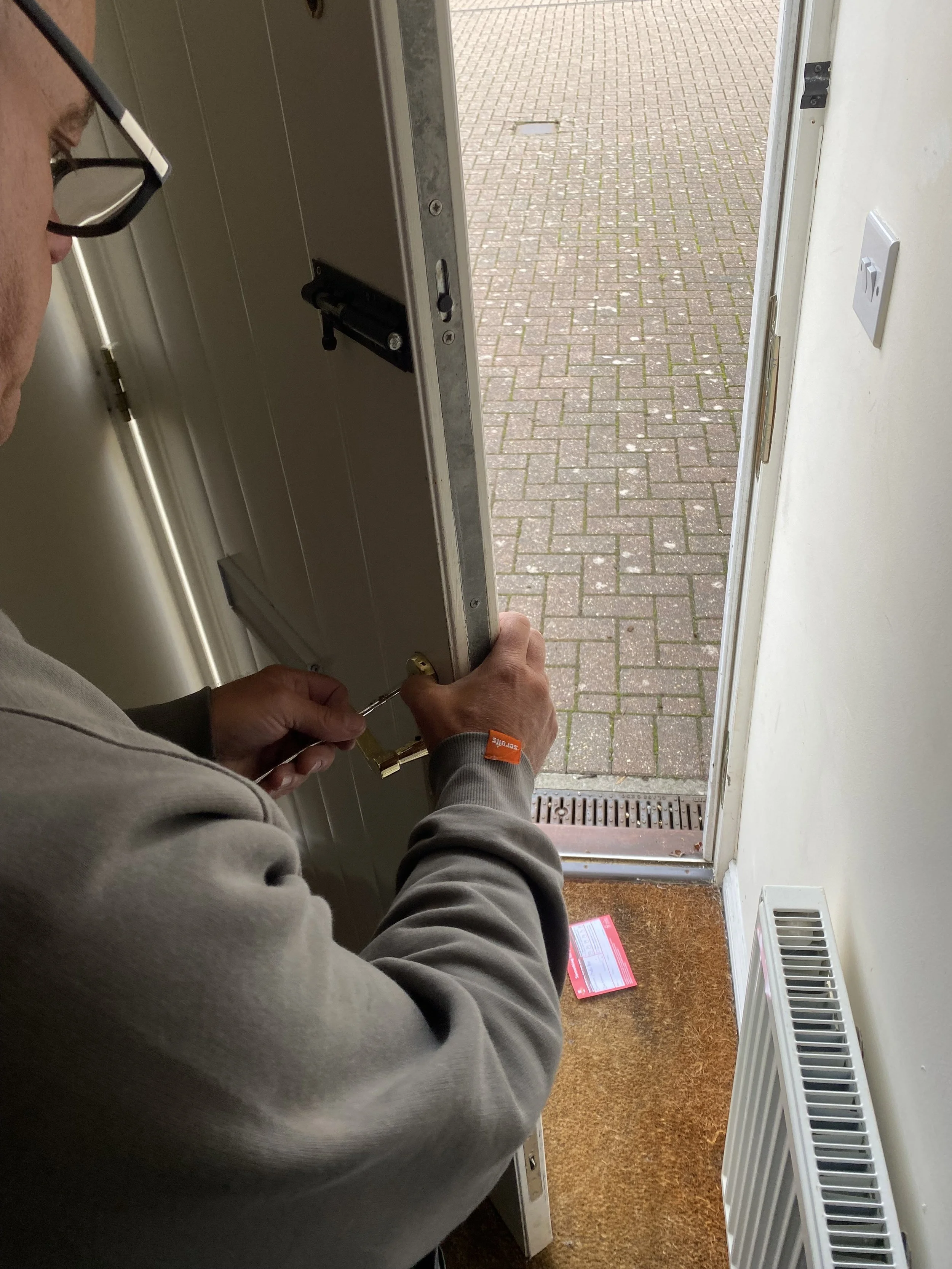 Person installing a lock on a door inside a building, with a brick-paved outdoor area visible through the open door.
