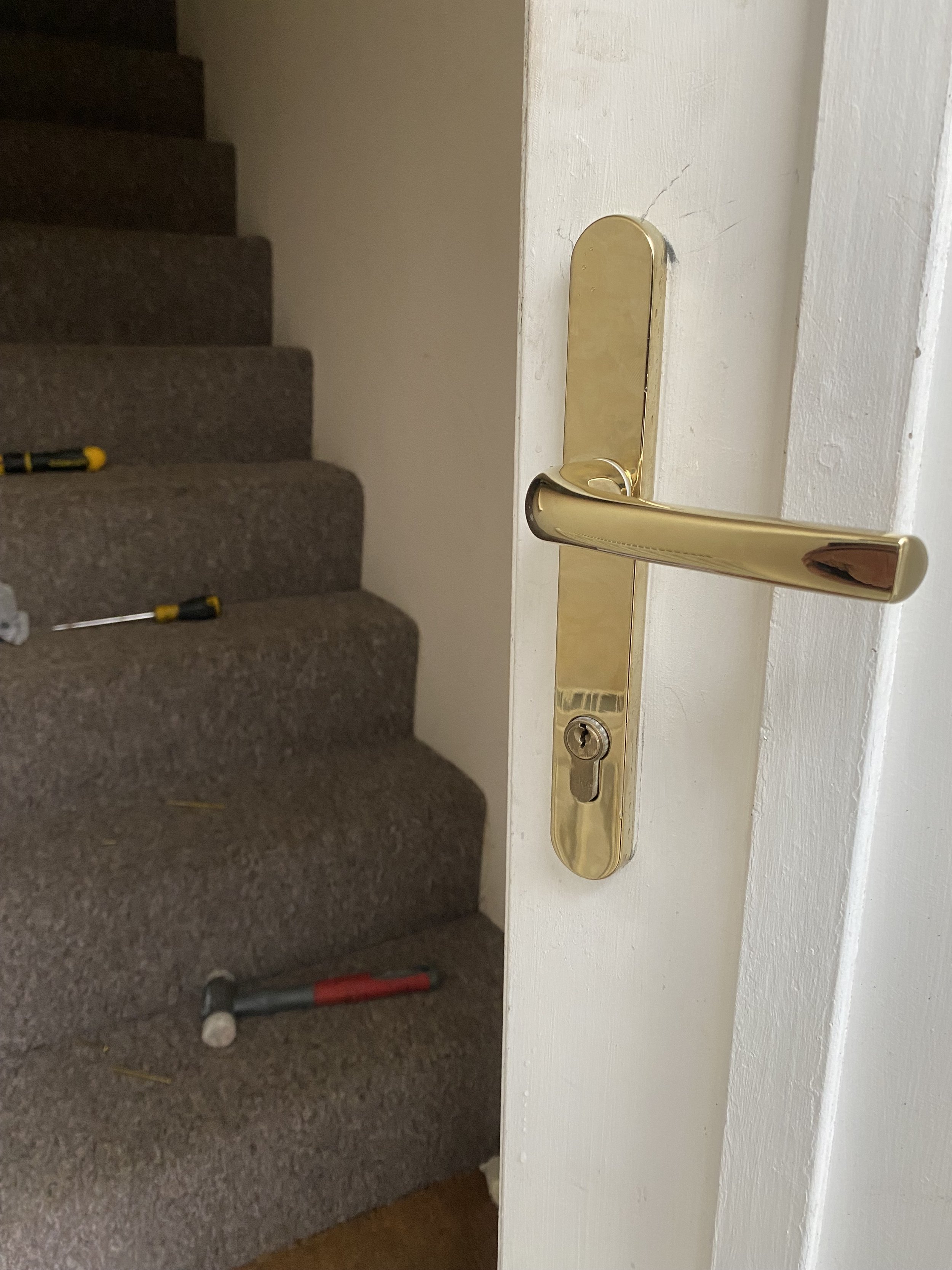 A partially open white door with a gold handle and lock, revealing a staircase with brown carpeted steps and tools such as a hammer, screwdriver, and screws on the stairs.