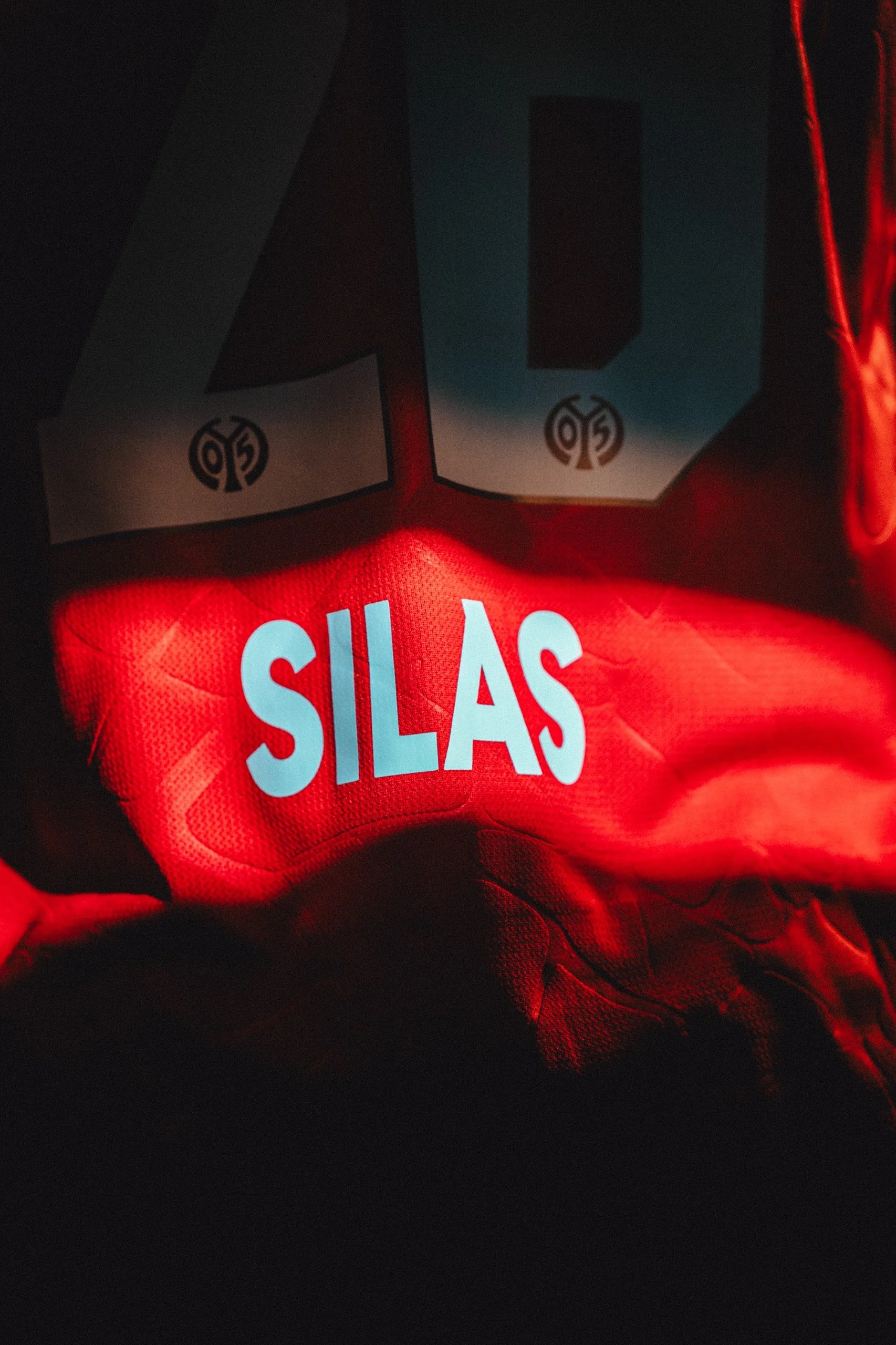 Close-up of a sports jersey with the name 'SILAS' printed in white, under a bold, blue number 0 on a dark background, illuminated by red light.