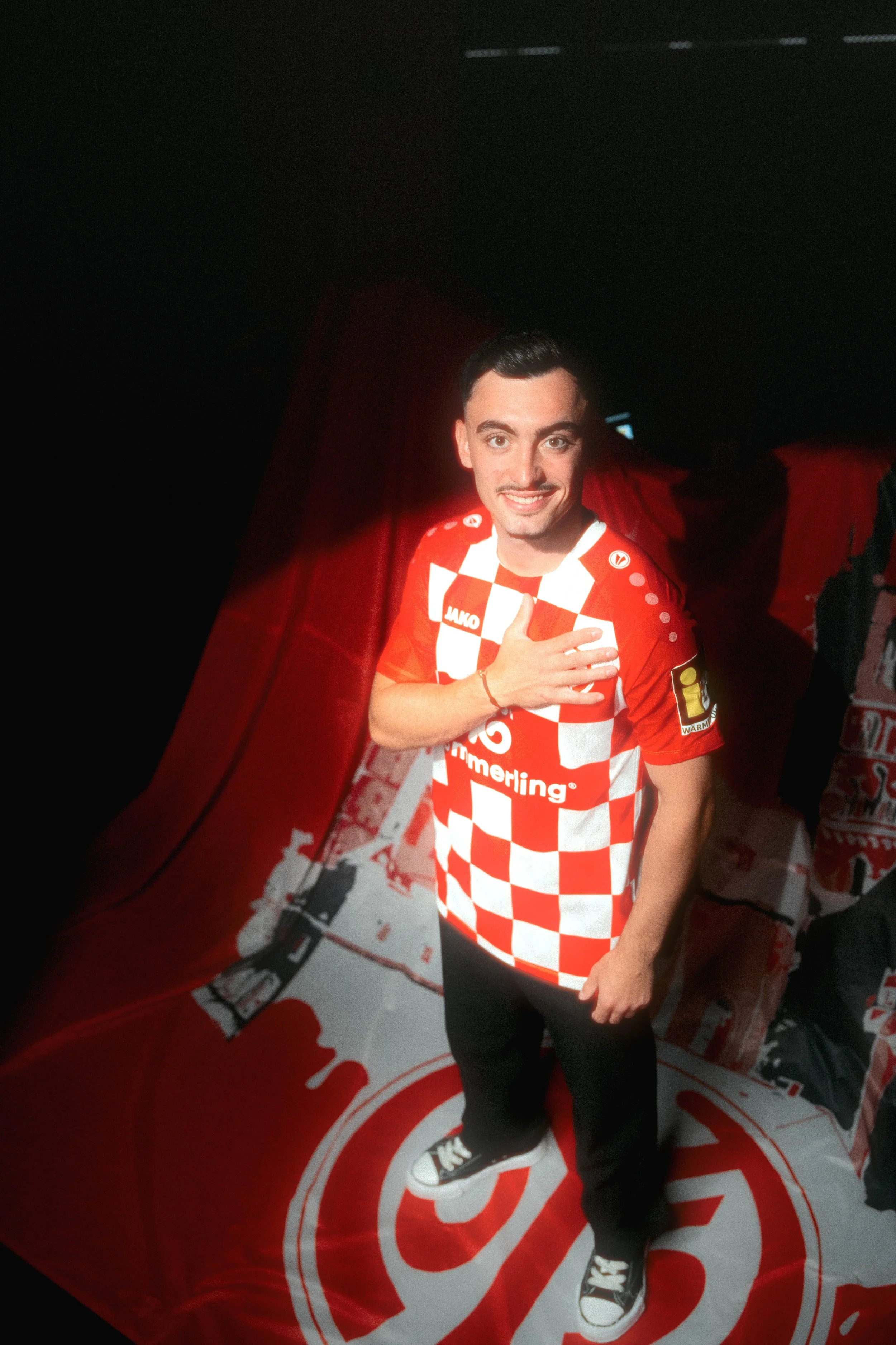 A young man with dark hair and light skin wearing a red and white checkered soccer jersey, black pants, and black sneakers with white soles, standing on a red and white floor with a large logo. He is smiling, looking at the camera, and has one hand o