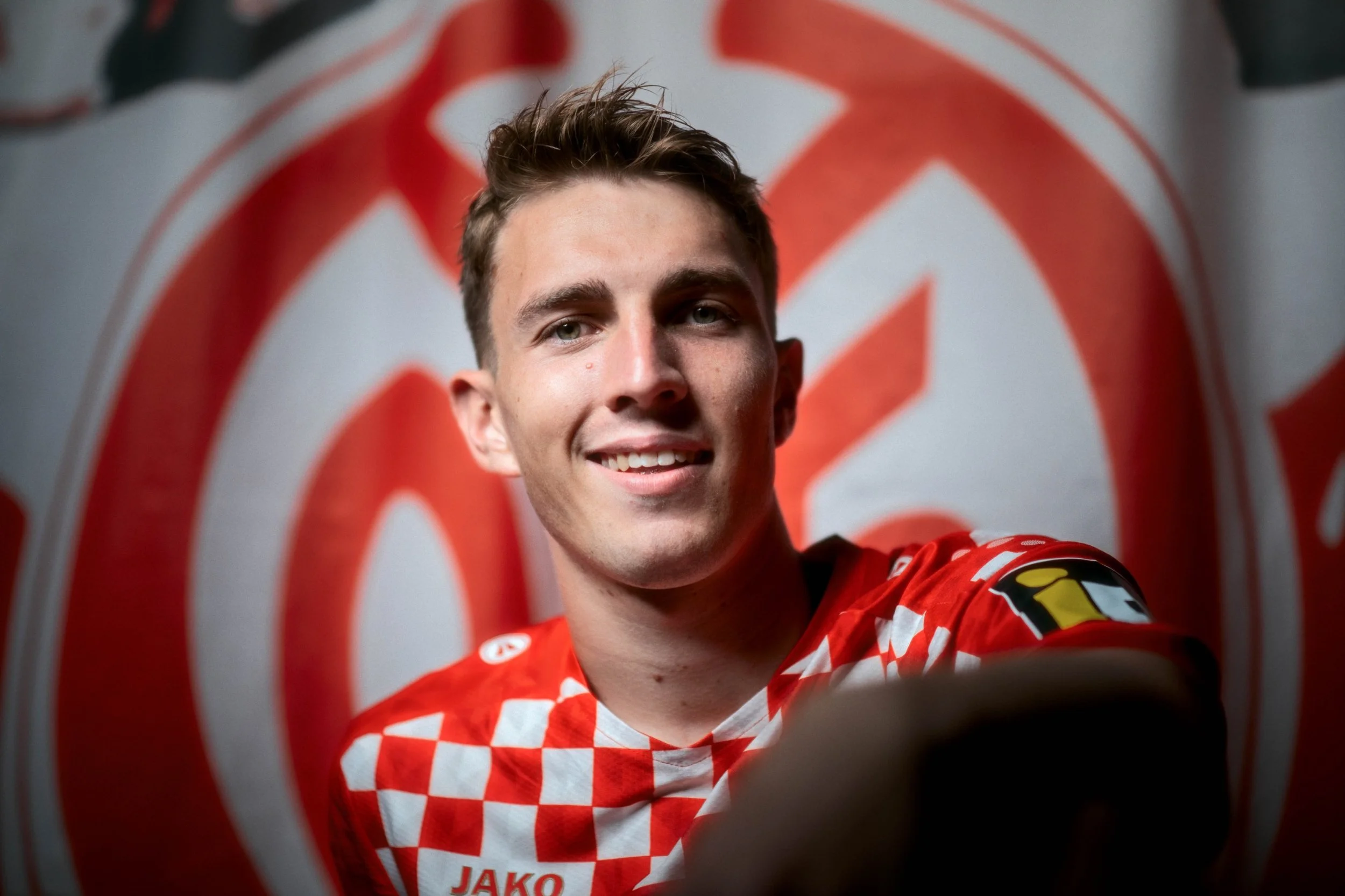A young man smiling in front of a winnipeg jets logo, wearing a red checkered hockey jersey.