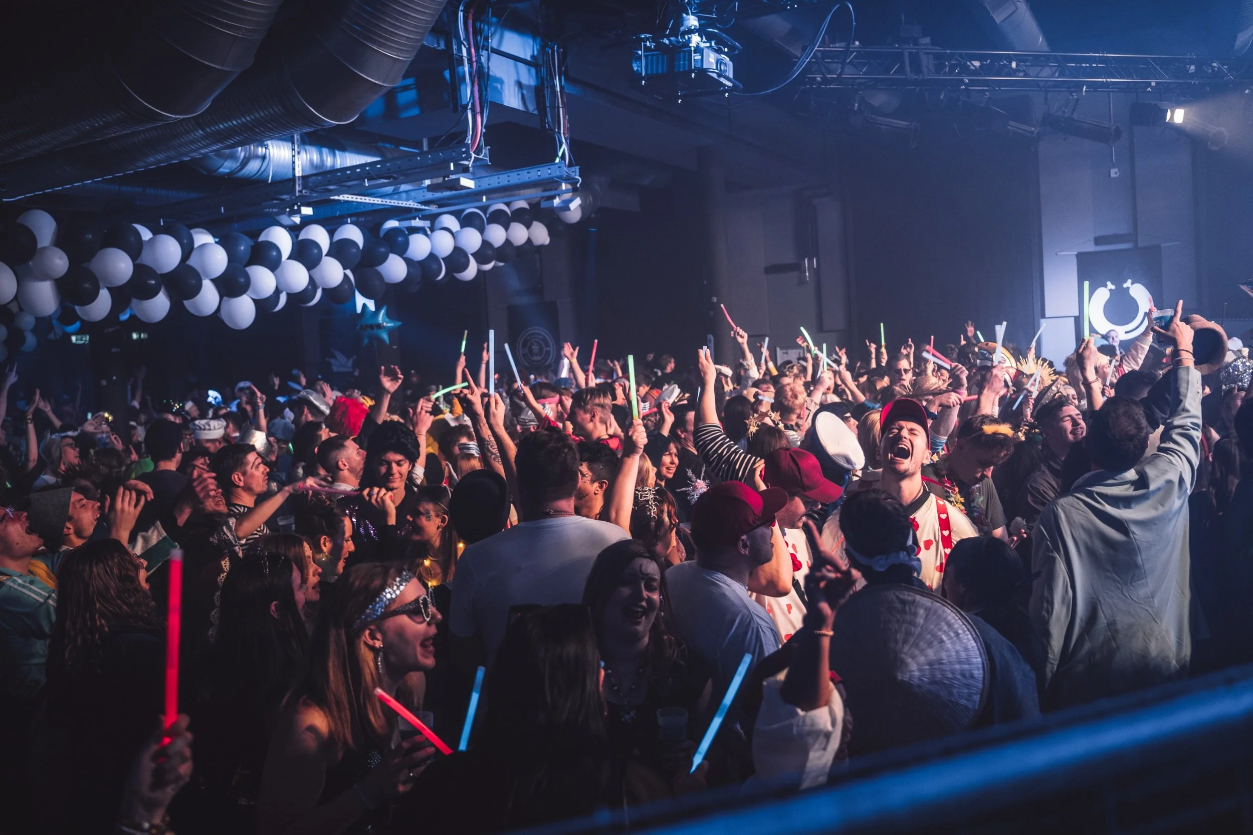 Crowd of people at an indoor concert or party, dancing, holding glow sticks, with black and white balloons and stage lighting in the background.