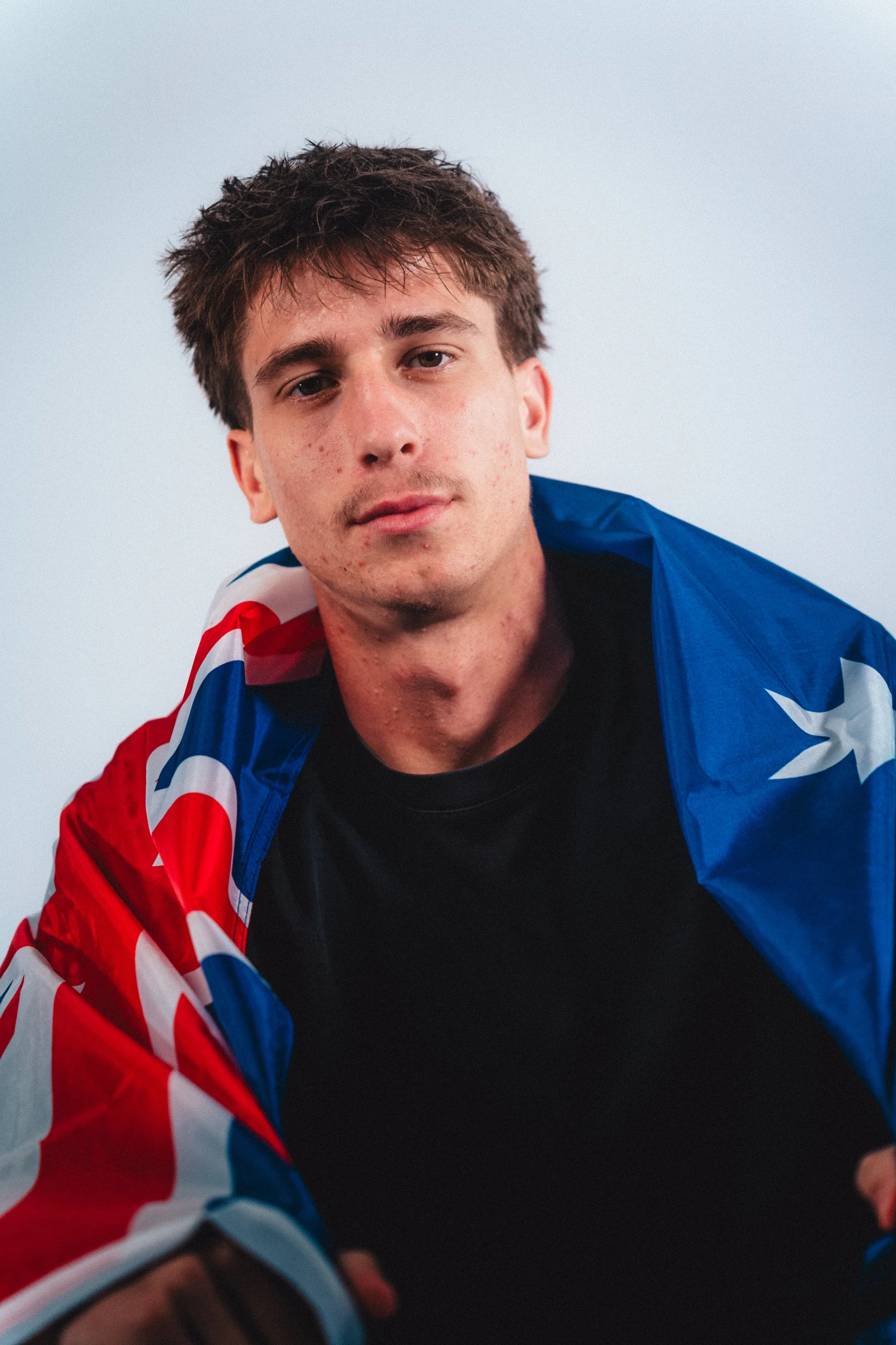 Young man with brown hair and acne, looking at the camera, wearing a black shirt and draped with the Australian flag.
