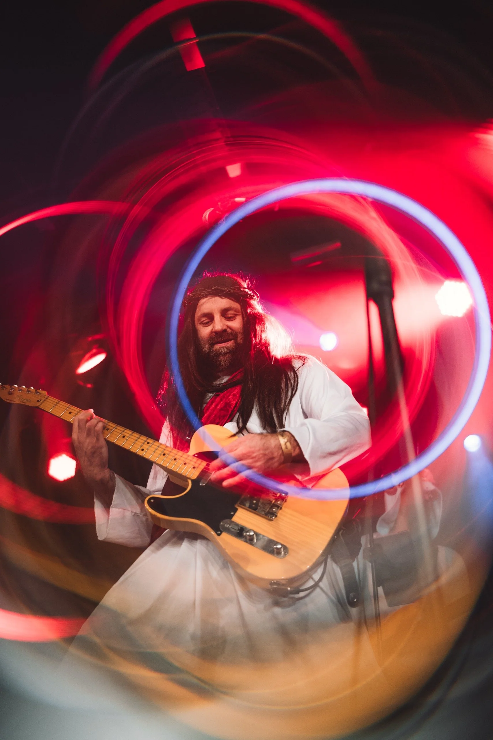 Musician with long hair playing an electric guitar on stage, surrounded by colorful light trails.