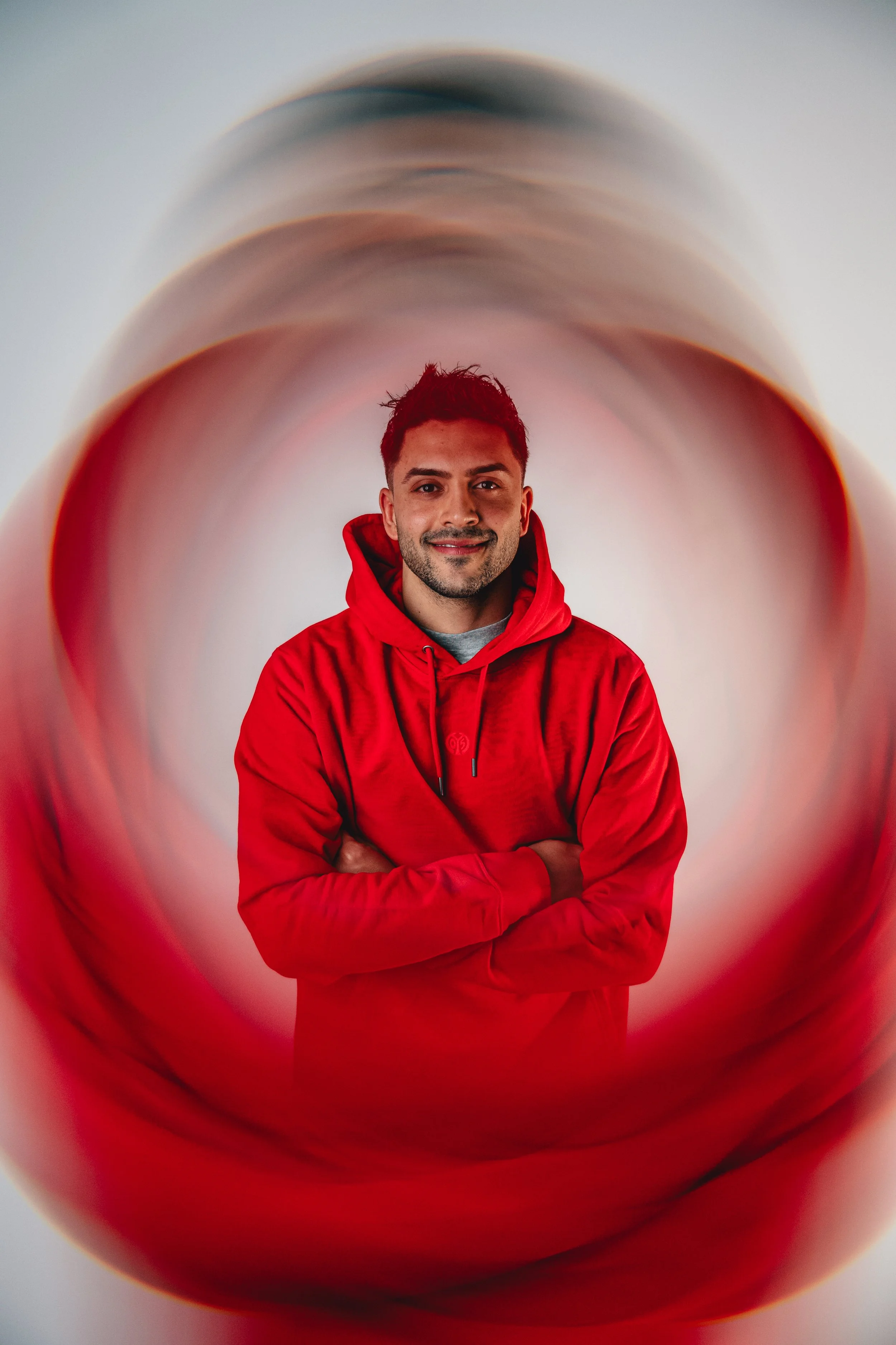 A man in a red hoodie standing inside a large, hollow, red-colored pipe or tube, looking at the camera and smiling.