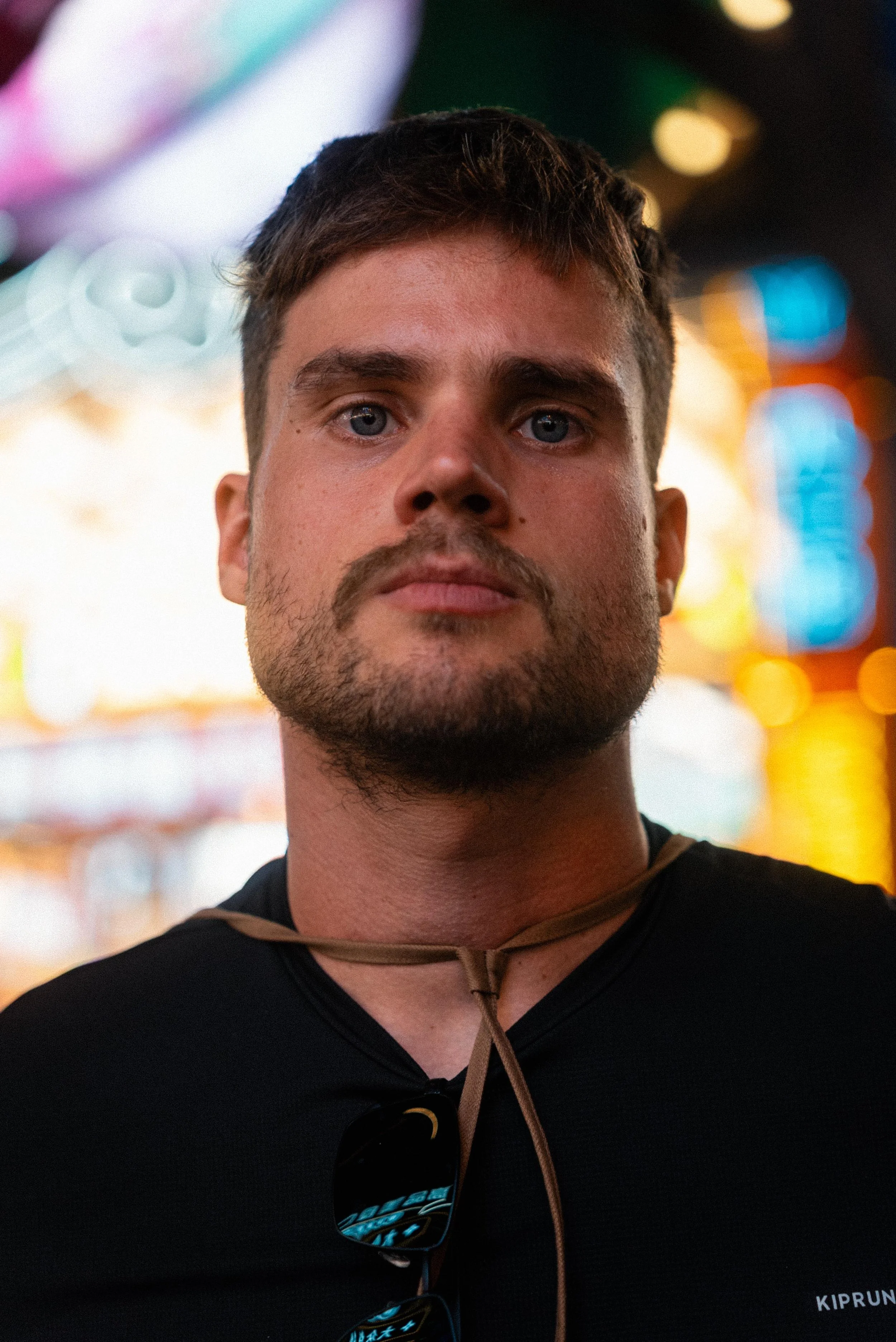 Close-up portrait of a man with short brown hair, light skin, and blue eyes, wearing a black shirt with a logo and sunglasses hanging from his shirt, standing in front of colorful bright lights in a night setting.