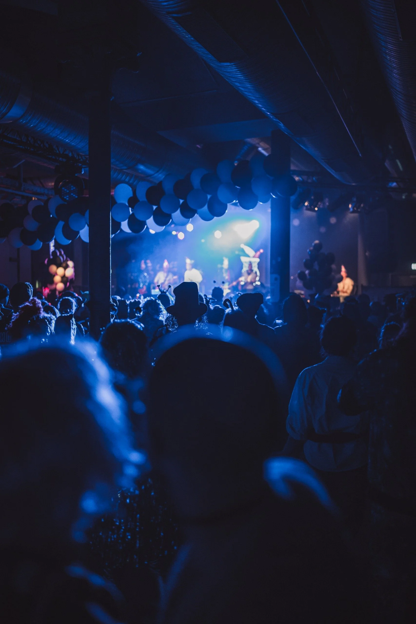 Crowd watching a stage performance in a dark, decorated venue with balloons and blue lighting.