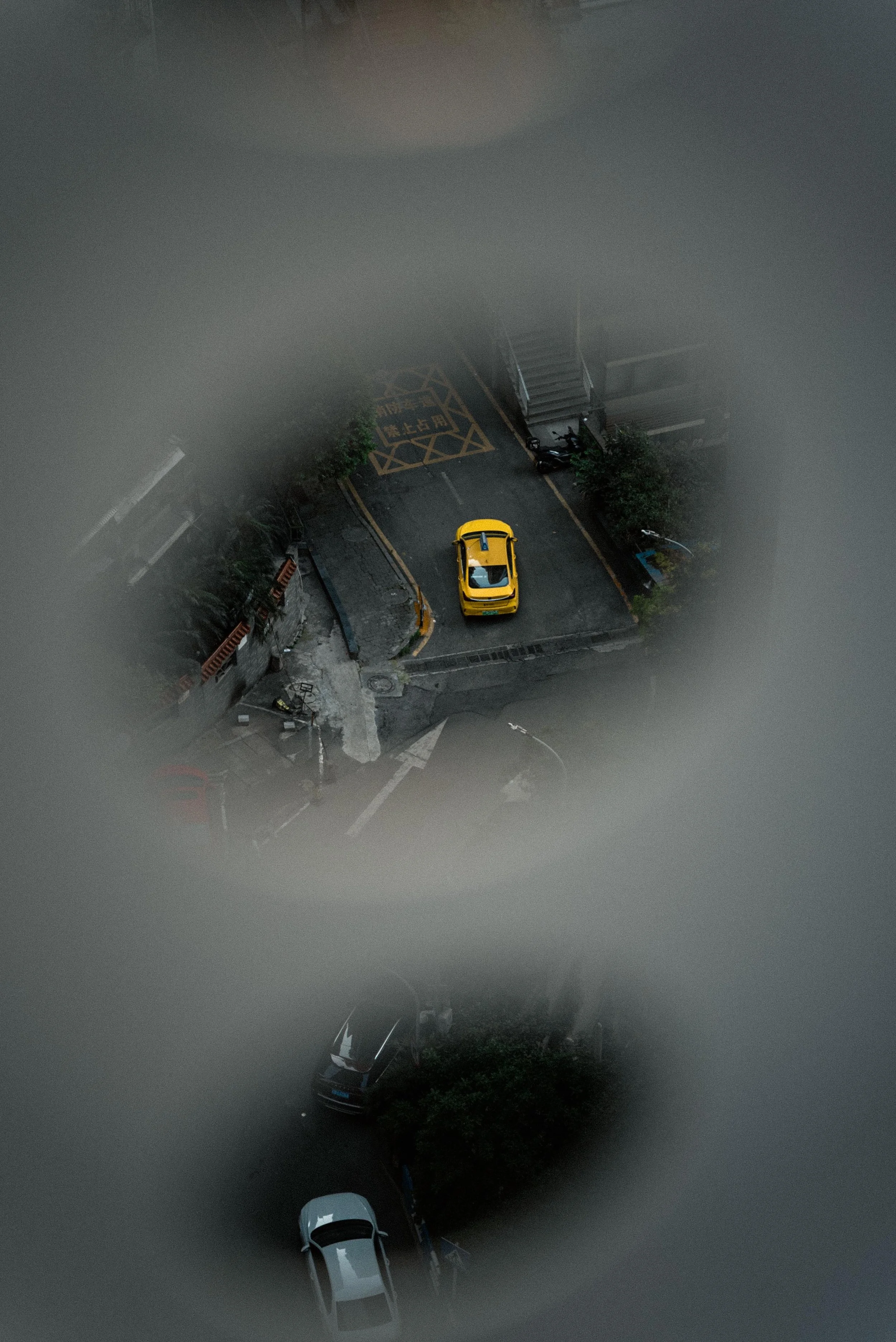View of a city street seen from a high vantage point through a circular opening, showing a yellow taxi on the road with other parked cars and buildings around.
