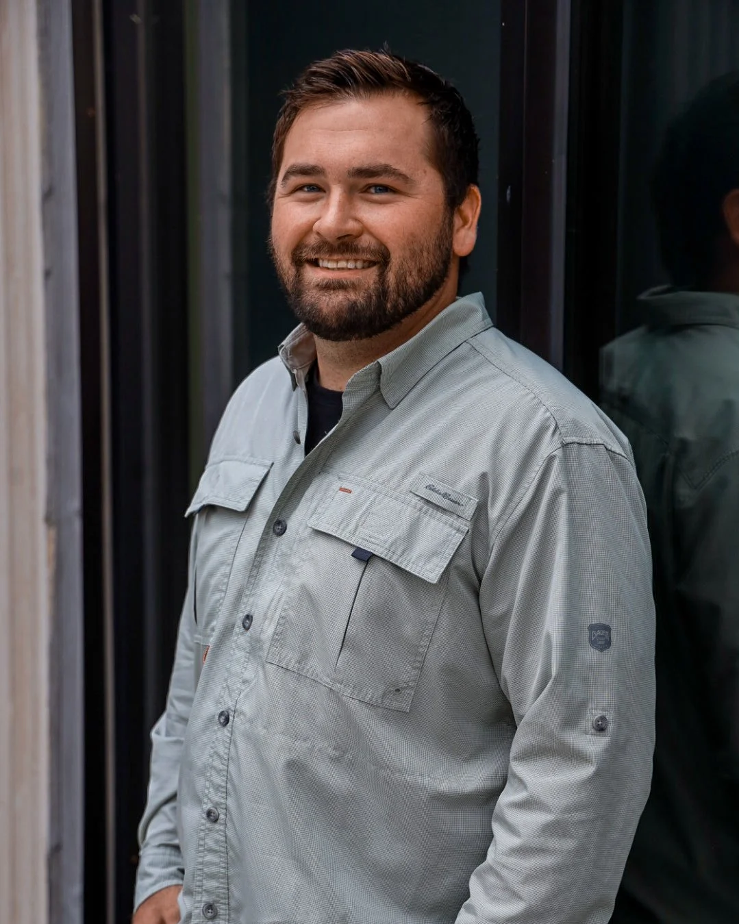 A man with a beard and short dark hair, smiling, wearing a light-colored button-up shirt, standing outdoors near a reflective glass wall.