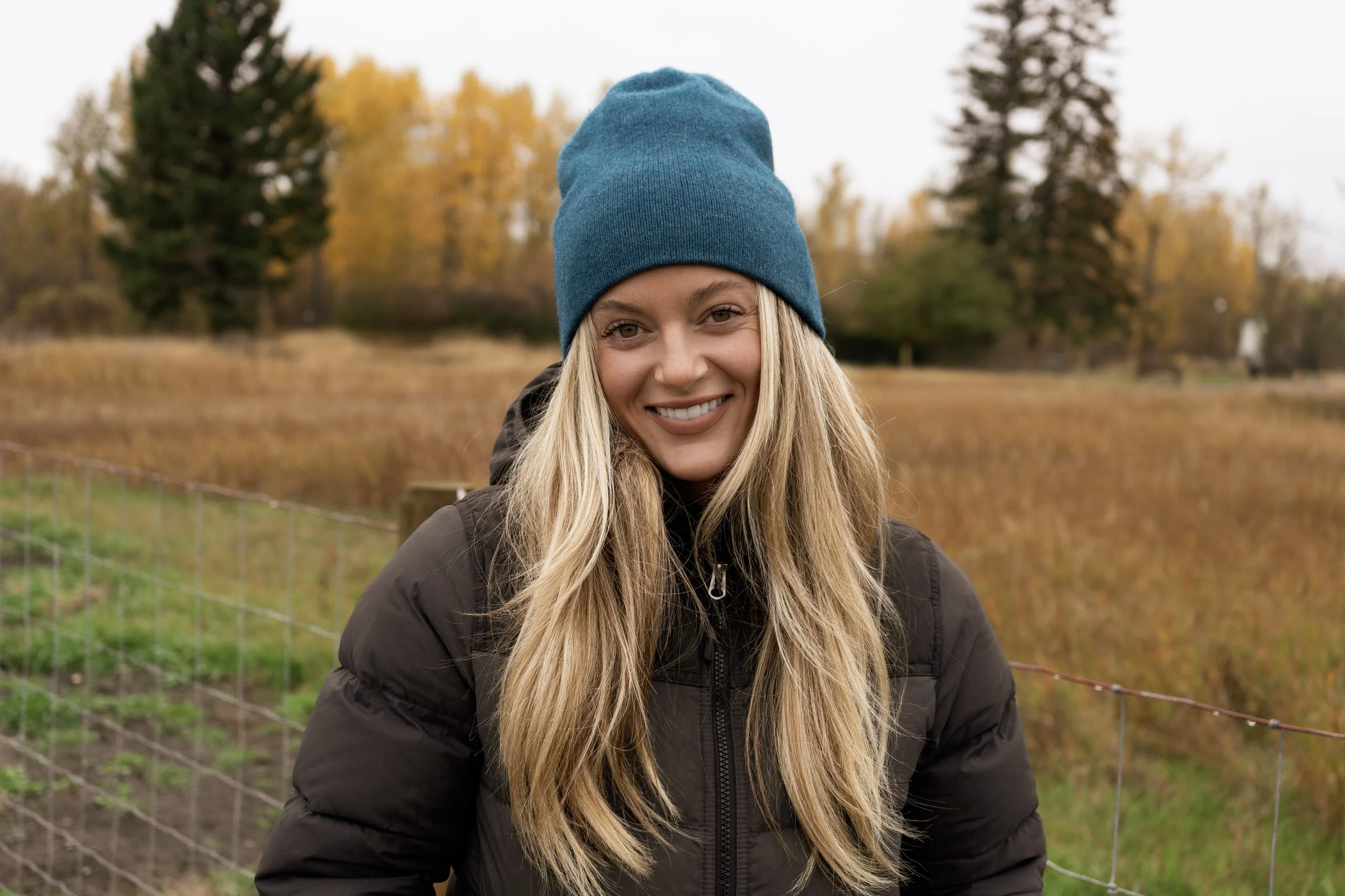A smiling woman with long blonde hair wearing a blue beanie and black jacket outdoors in fall, near a wire fence with trees and autumn foliage in the background.