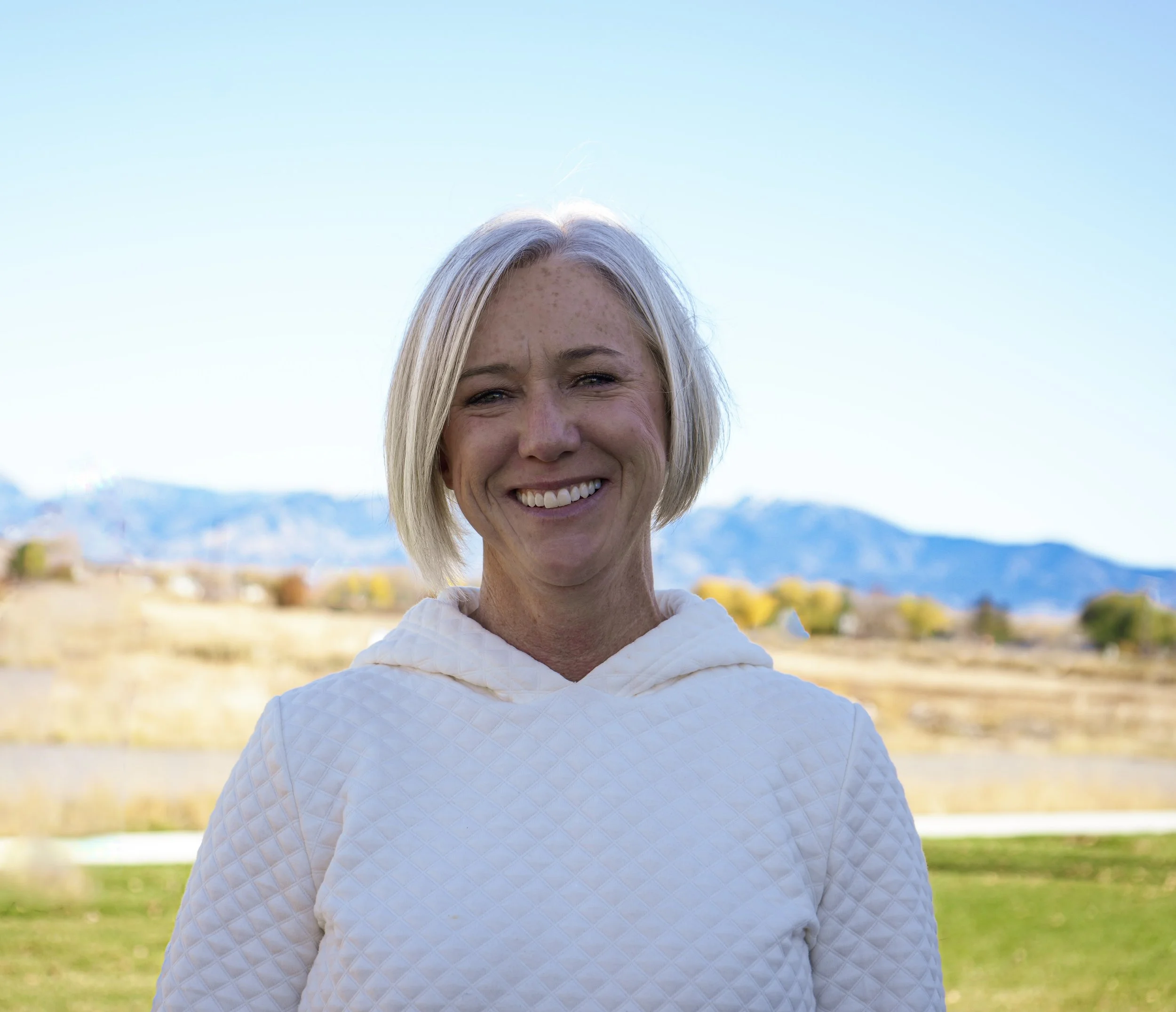 A smiling woman with short blonde hair standing outdoors with mountains in the background.