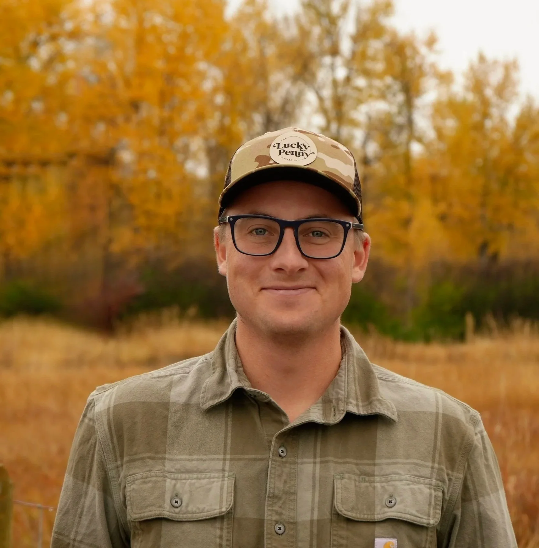 A man outdoors in fall, wearing glasses, a camouflage cap with 'Lucky Penny' logo, and a beige plaid shirt, standing in front of yellow-orange autumn trees.