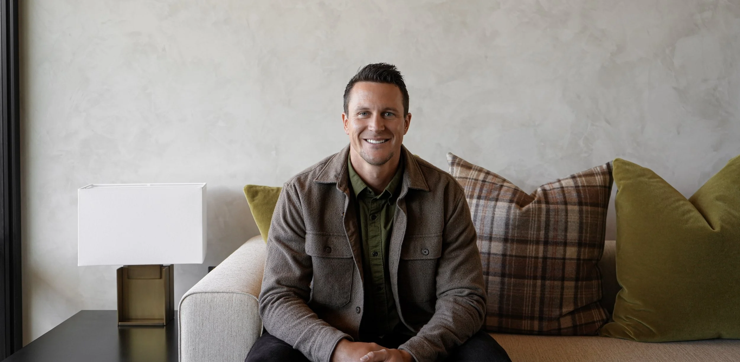 A smiling man with short dark hair, wearing a green shirt and brown jacket, sitting on a beige couch with green and brown plaid and plain green pillows in a modern living room.