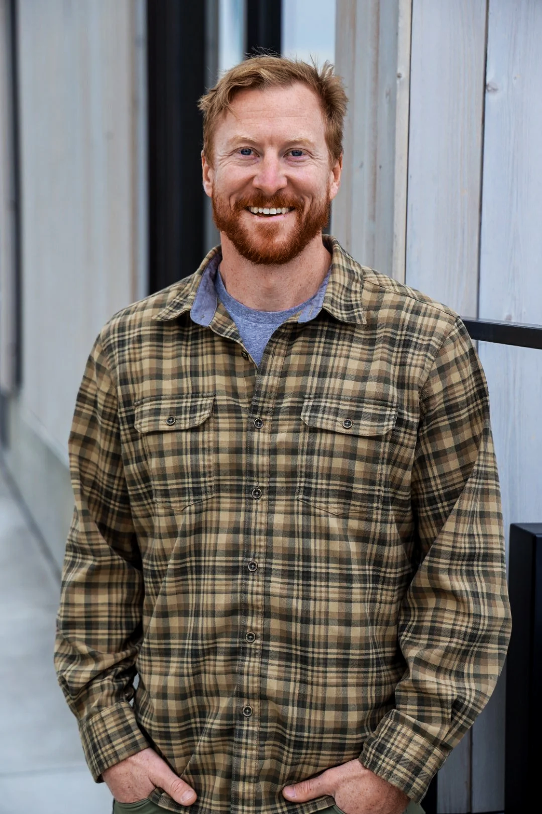 A woman with long brown hair, wearing a white blazer, smiles and looks at the camera, sitting on a tan leather couch in a bright room.