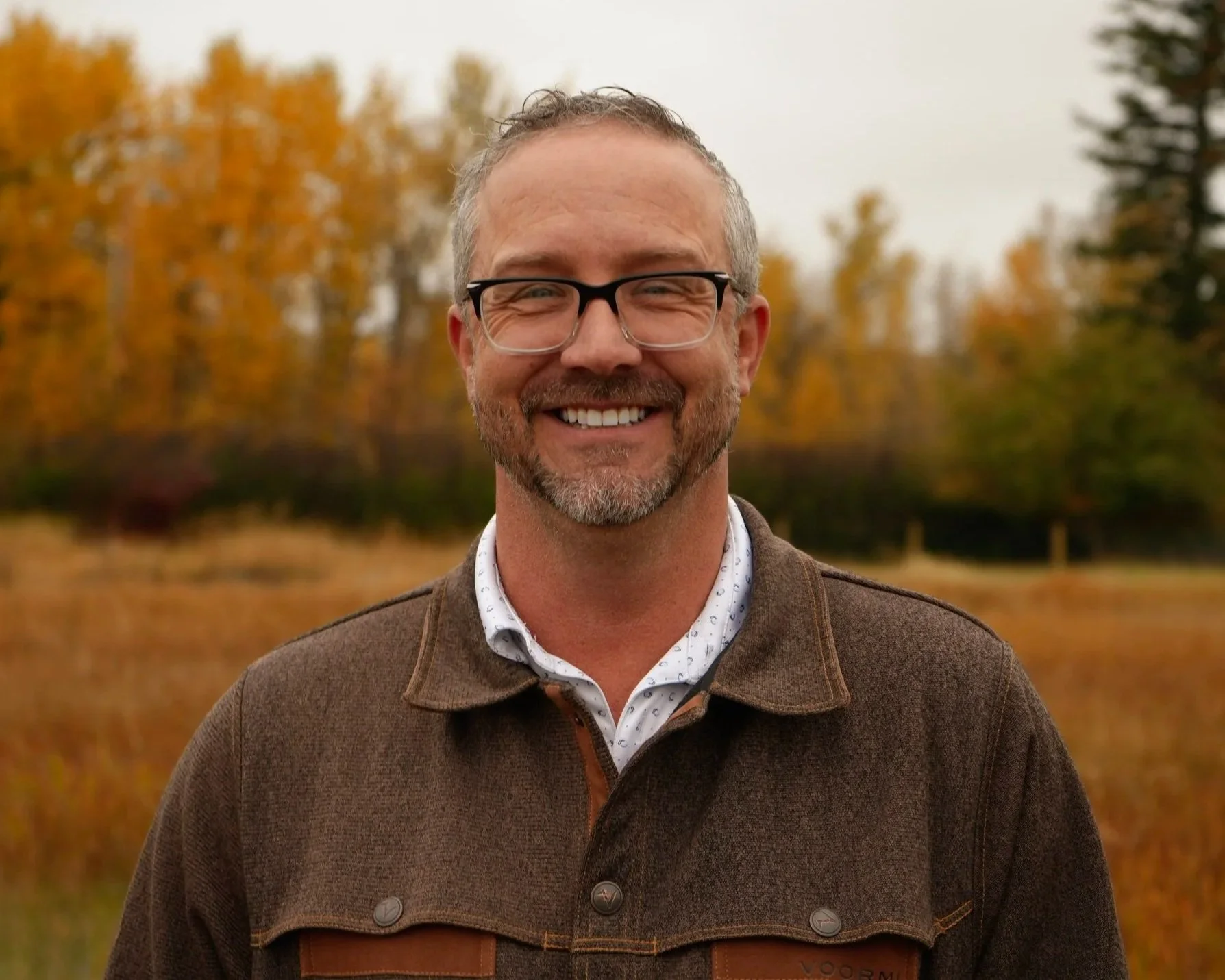 A smiling man with red hair and a beard, wearing a plaid shirt, standing outdoors near a building with wooden siding.